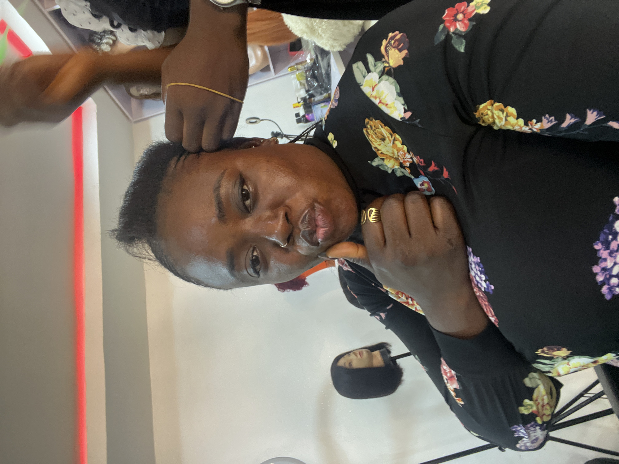 This image, reportedly captured in Abuja, Nigeria, depicts an adult Black woman receiving a hair treatment in what appears to be a salon setting. The woman occupies the central foreground, looking directly at the camera with pursed lips. Her right hand is raised in a closed-fist gesture, with a gold-toned ring visible on her ring finger. She wears a long-sleeved black top adorned with a vibrant floral pattern featuring yellow, red, purple, and white blossoms. Her dark hair is being actively styled; the hands of a second individual are visible in the upper right, manipulating strands near the woman's left temple, consistent with braiding or twisting. A thin gold elastic band and a silver-toned wristwatch are visible on the stylist's left wrist.
The background reveals typical salon elements. To the left, a dark bob-style wig is displayed on a mannequin head atop a stand. A portion of a ring light apparatus is also visible on the far left. In the upper right background, shelves are stocked with various hair products, including bottles and items enclosed in clear packaging. Another wig head, featuring a lighter-colored wig, is partially visible on these shelves. A prominent red linear light fixture is affixed to the ceiling in the upper background. The walls of the establishment are light-colored.