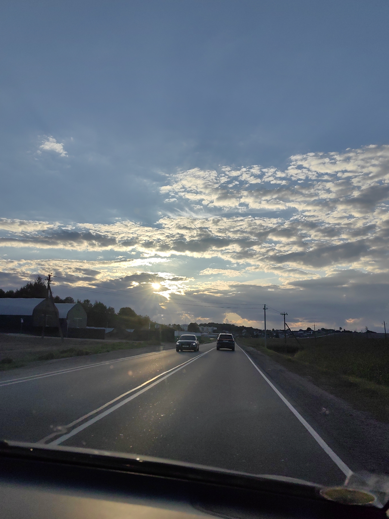 The image shows a view from inside a vehicle traveling on a road. Two cars are visible ahead on the same road, both heading in the same direction. The sky is filled with a mix of blue sky and scattered clouds, with the sun breaking through the clouds, casting rays of light. The landscape on either side of the road includes fields, trees, and some buildings, possibly farm structures. There are utility poles with wires along the side of the road. The scene appears to be in a rural or semi-rural area. Based on the information provided, the location is Dzyarzhynsk, Belarus. The time of day appears to be late afternoon or early evening due to the angle of the sun and the lighting conditions.
