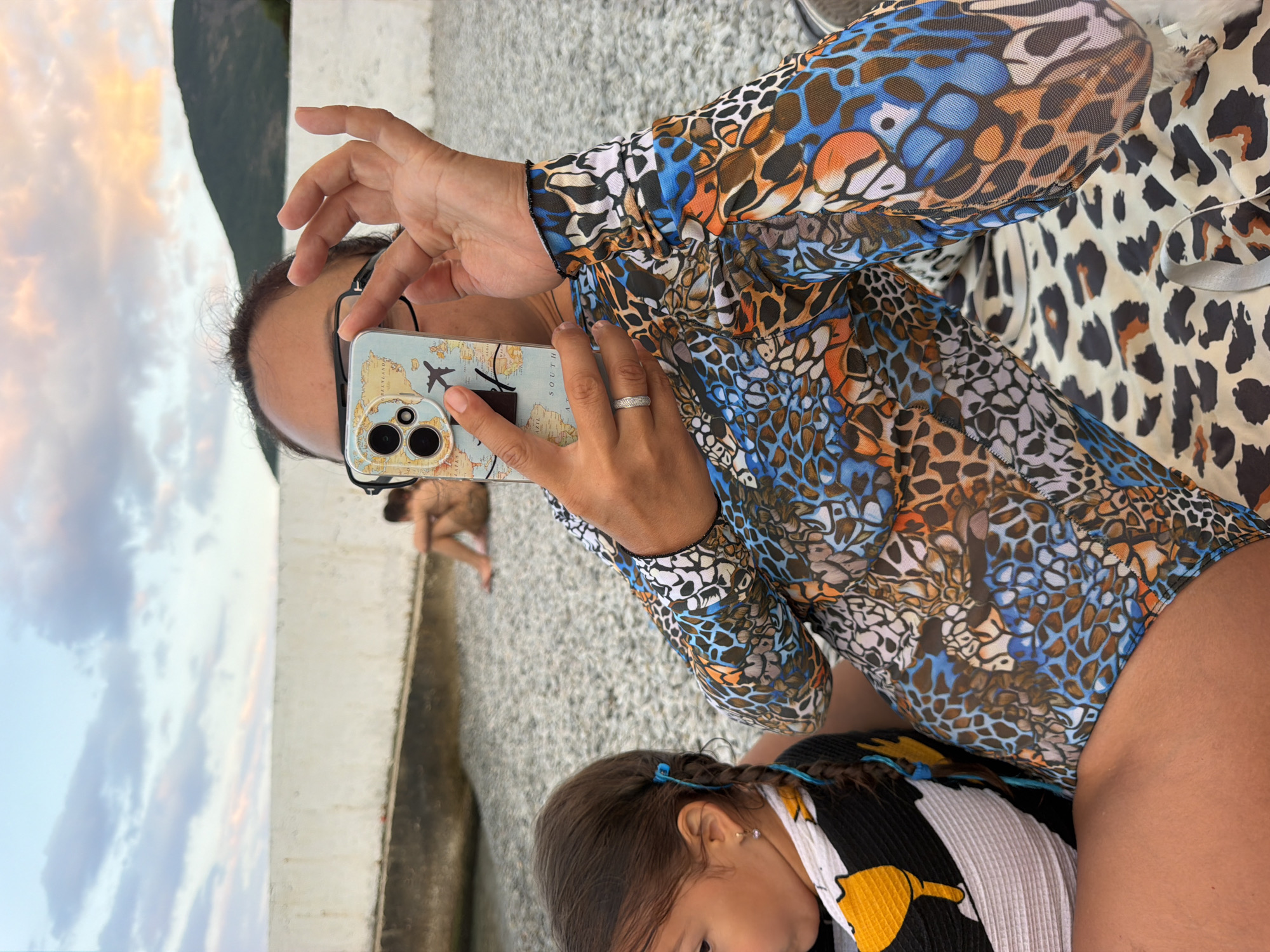 An image taken at a beach in Partenit, Ukraine shows a woman holding a phone up to take a photo. She is wearing a brightly colored, patterned swimsuit with long sleeves. In the foreground, a young child is visible, with their face turned away from the camera. The background shows a rocky beach area, a white wall, and a hill in the distance under a cloudy sky. Another person is seated in the background. The overall scene depicts a relaxed outdoor setting by the sea.
