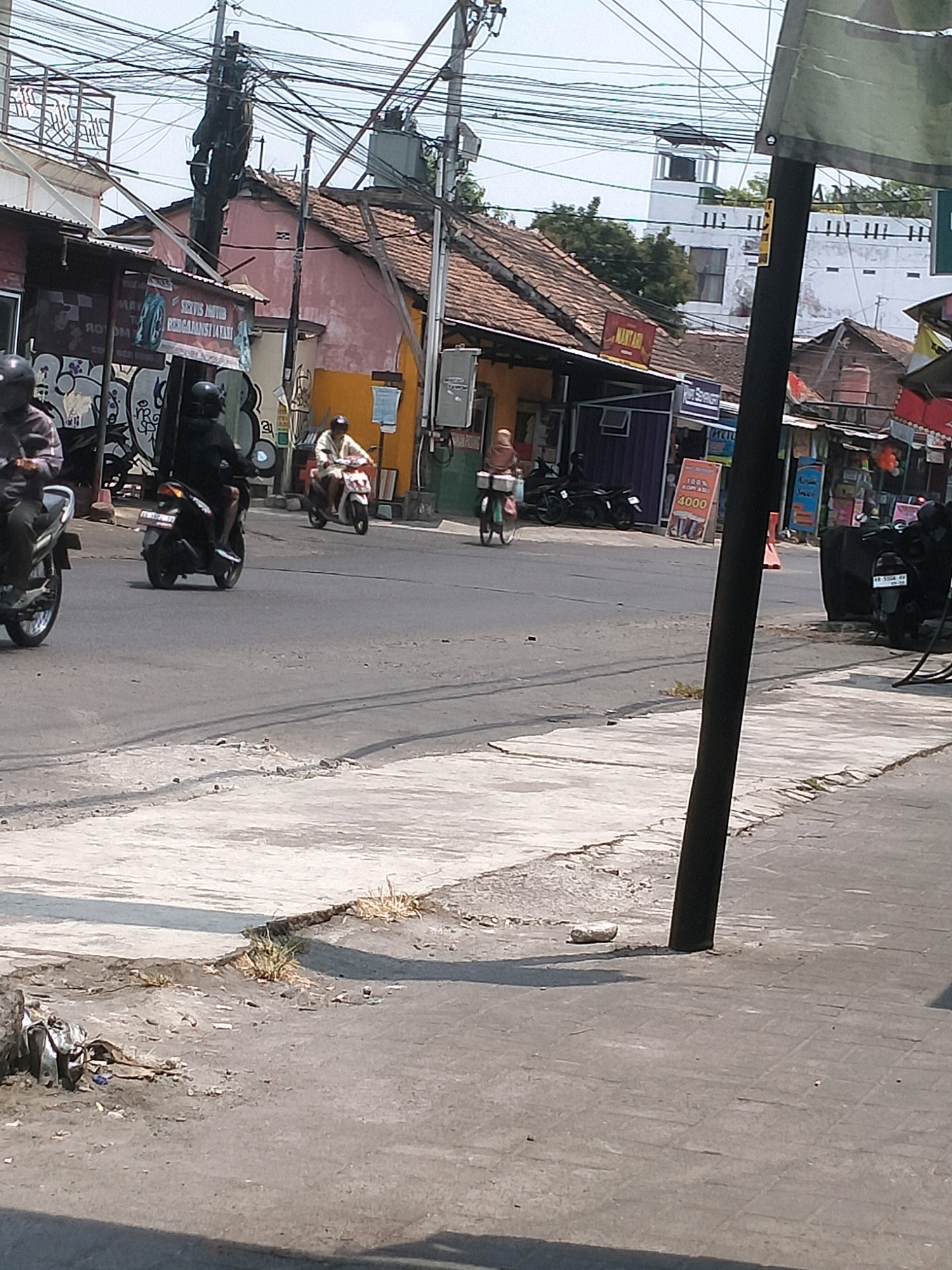The image depicts a street scene in Kasihan, Indonesia. In the foreground, a paved sidewalk runs along the edge of a road. The road surface is asphalt and shows signs of wear, with some cracks and patches. Several motorcycles are visible on the road. One motorcycle, ridden by a person wearing a black helmet and dark clothing, is in the left foreground, partially cut off by the frame. Further down the road, another motorcycle is ridden by a person in black with a helmet. In the middle ground, a person on a white and red motorcycle is seen. A person on a bicycle with a basket is also visible further down the road. The background features buildings with red-tiled roofs and some with brightly colored walls. Overhead, a complex network of electrical wires and poles dominates the sky. Signs are visible on the buildings, some with text in Indonesian. Vegetation is sparse, with some small bushes growing on the roadside. The overall impression is a busy urban street with traffic and commercial activity.
