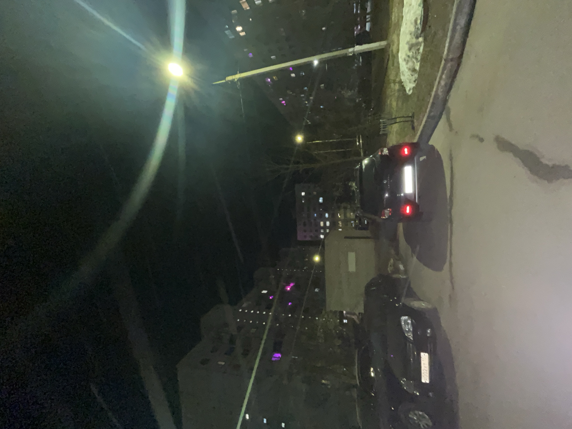 The image shows a street scene at night in what appears to be Klin, Russia. A streetlamp illuminates the foreground. Two black cars are parked on the side of a road. A multi-story building with lit windows is visible in the background.