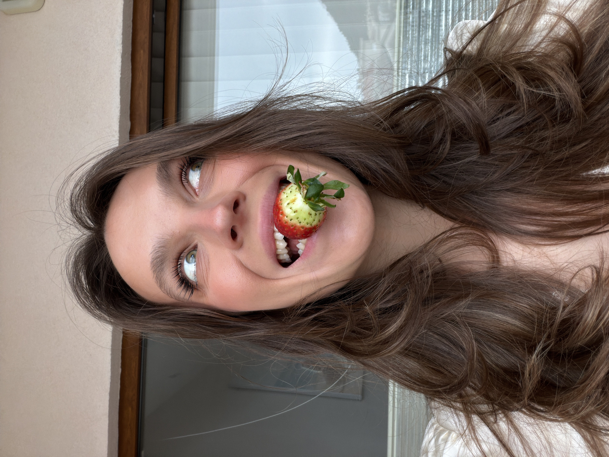 A close-up, chest-up image depicts a female subject with long, wavy brown hair, fair complexion, and green eyes. Her mouth is open in a wide smile, revealing white teeth, as she holds a strawberry between her upper and lower front teeth. The strawberry displays red ripeness on one side and a light green/white hue on the other, with green sepals and a short stem visible. Her eyes are directed upwards and slightly to the right. She is wearing a light-colored garment, possibly white, with gathered fabric visible on her shoulders.
The background consists of an interior wall section in light beige or off-white, situated above a dark brown wooden window or door frame. Beyond this frame, an exterior scene is visible through glass. This exterior features an overcast, light grey sky and distant, hazy mountains or hills. Faint architectural elements are discernible below the mountain line. The scene is consistent with a balcony view in the city of Budva, Montenegro.