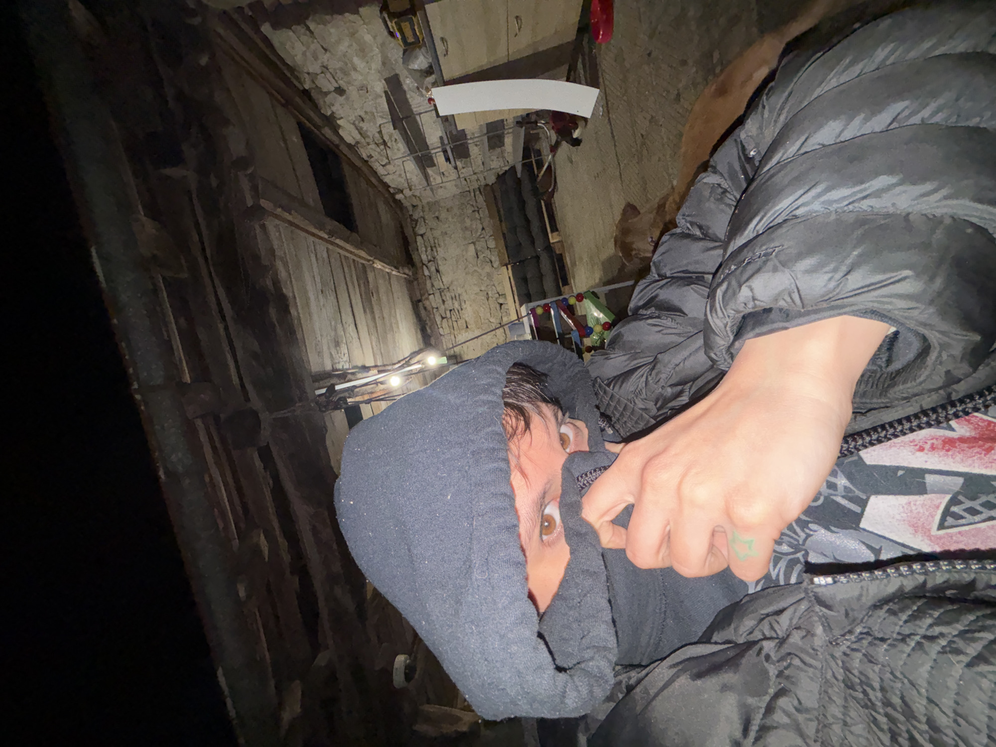 The image shows a person in a gray hooded sweatshirt and a black puffer jacket, with their face partially obscured by the hood. Their hand is near their face. The location appears to be an alleyway in the village of Karlukovo, Bulgaria, judging by the presence of traditional stone and wood buildings. The alley is narrow and lit by dim overhead lights.