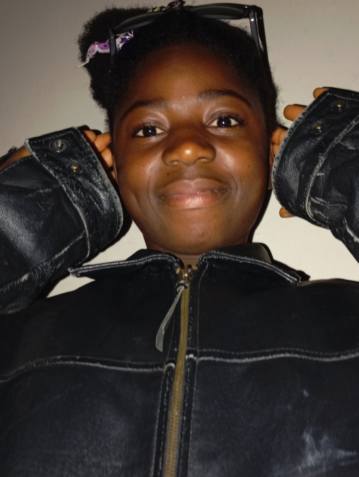 A close-up shot of a young, dark-skinned person wearing a black leather jacket. The person is holding the flaps of the jacket near their ears. Sunglasses rest on top of their head, and a patterned hair accessory is visible. The background is a plain, light-colored wall. The location in the city of Jos, Nigeria is not discernible from the image.