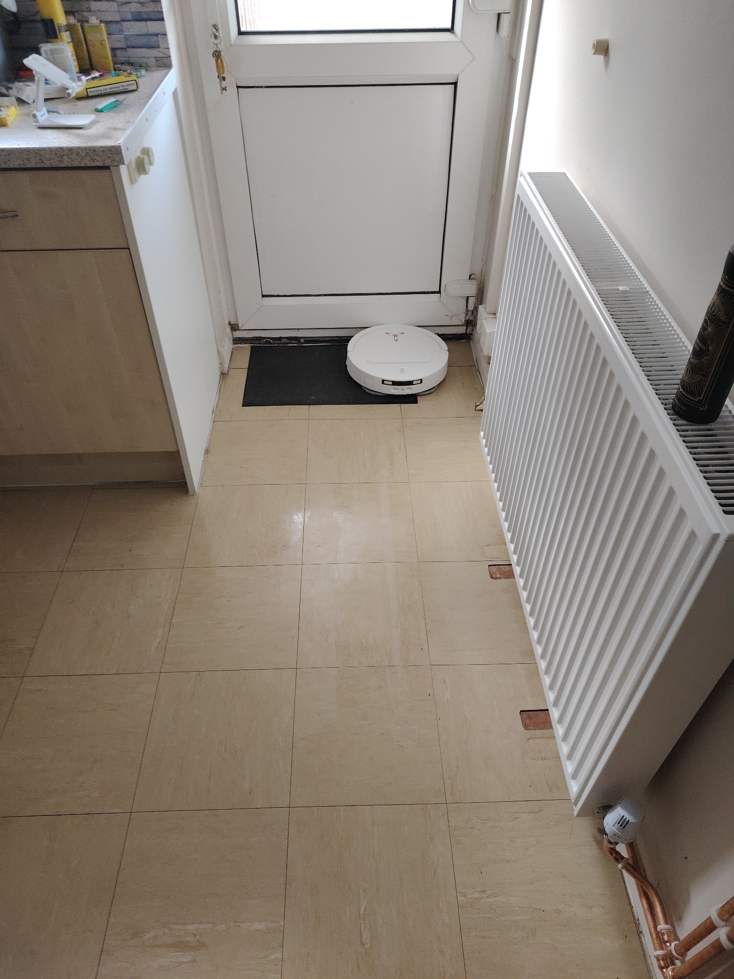This image displays an interior ground-level hallway or utility area, situated in Huntingdonshire, United Kingdom. The scene is lit by natural light originating from a window integrated into a door.
On the left side, a section of a light-colored wooden base cabinet with a speckled grey countertop is visible. The countertop supports two yellow cylindrical containers, a white mobile device stand, a green utensil or tool, and a white and red cigarette packet bearing a UK government health warning. Adjacent to the counter, a vertical white panel features two circular white stoppers.
On the right side of the frame, a white panel radiator is affixed to a light-colored wall. Beneath the radiator, uninsulated copper pipes, partially secured with white clips, connect to a white thermostatic radiator valve (TRV). A small, light-colored adhesive strip is visible on the wall above the radiator.
The floor is composed of light beige square tiles, featuring dark grout lines and signs of surface wear. Directly beneath the radiator, two rectangular sections of exposed reddish-brown subfloor material are evident, indicating damaged or absent tiles.
In the center, a white circular robot vacuum cleaner, marked with a gold-colored logo, is stationary on a rectangular black doormat. This doormat is positioned immediately in front of a white uPVC door, which comprises a solid lower panel and an upper window panel. A set of keys hangs from a metal hook on the white door frame. The skirting boards and the base of the door frame show dirt and grime accumulation. No individuals are present in the image.