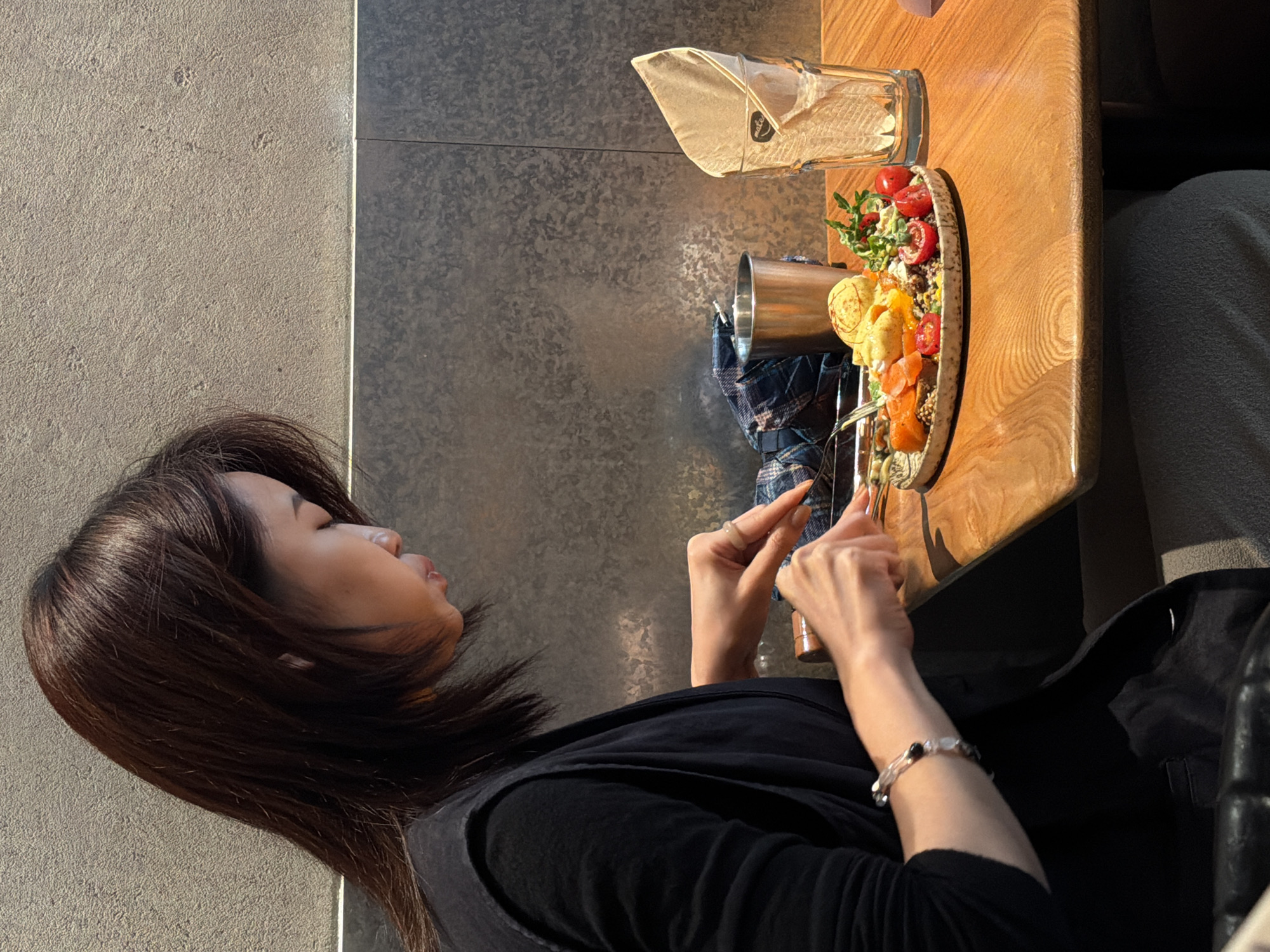 An Asian woman is seated at a wooden table in a restaurant, seen from the side. She is wearing a black vest over a black top, and a bracelet adorns her left wrist. The woman is focused on eating, using a fork and knife to cut into a plate of food. The plate contains a variety of items, including what appears to be eggs, salmon, tomatoes, and greens. To the right of the plate are a tall glass with a folded napkin inside and a metal cup. The background consists of a textured, gray wall and a dark gray lower wall. The restaurant is located in Yuzhong District, China.