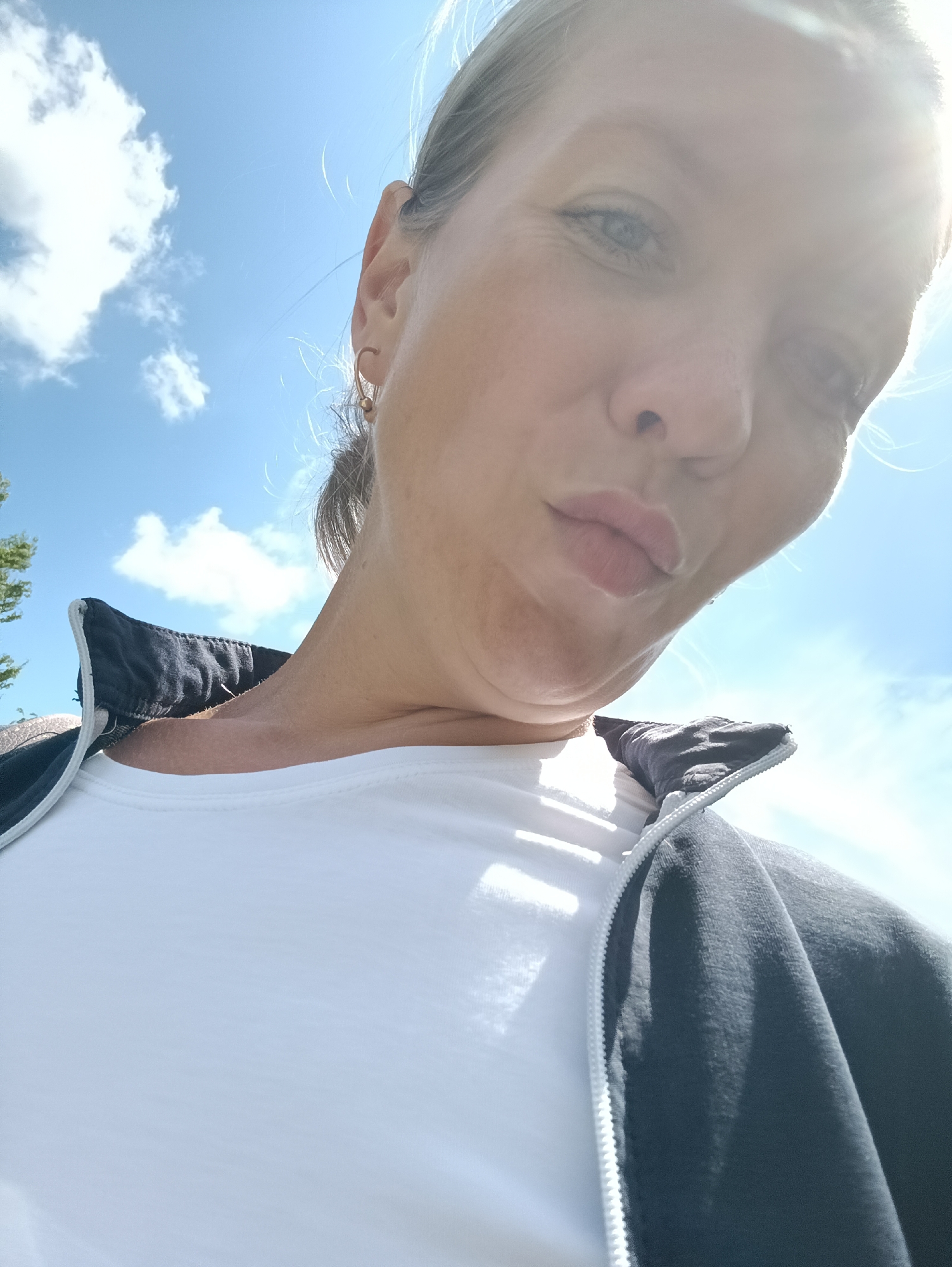 A woman wearing a white t-shirt and a black jacket is taking a selfie outdoors. She is looking down at the camera, with her lips pursed. The background is a bright blue sky with white clouds. A tree is visible on the left side of the image. The lighting suggests it is a sunny day. The woman is wearing a small gold earring.