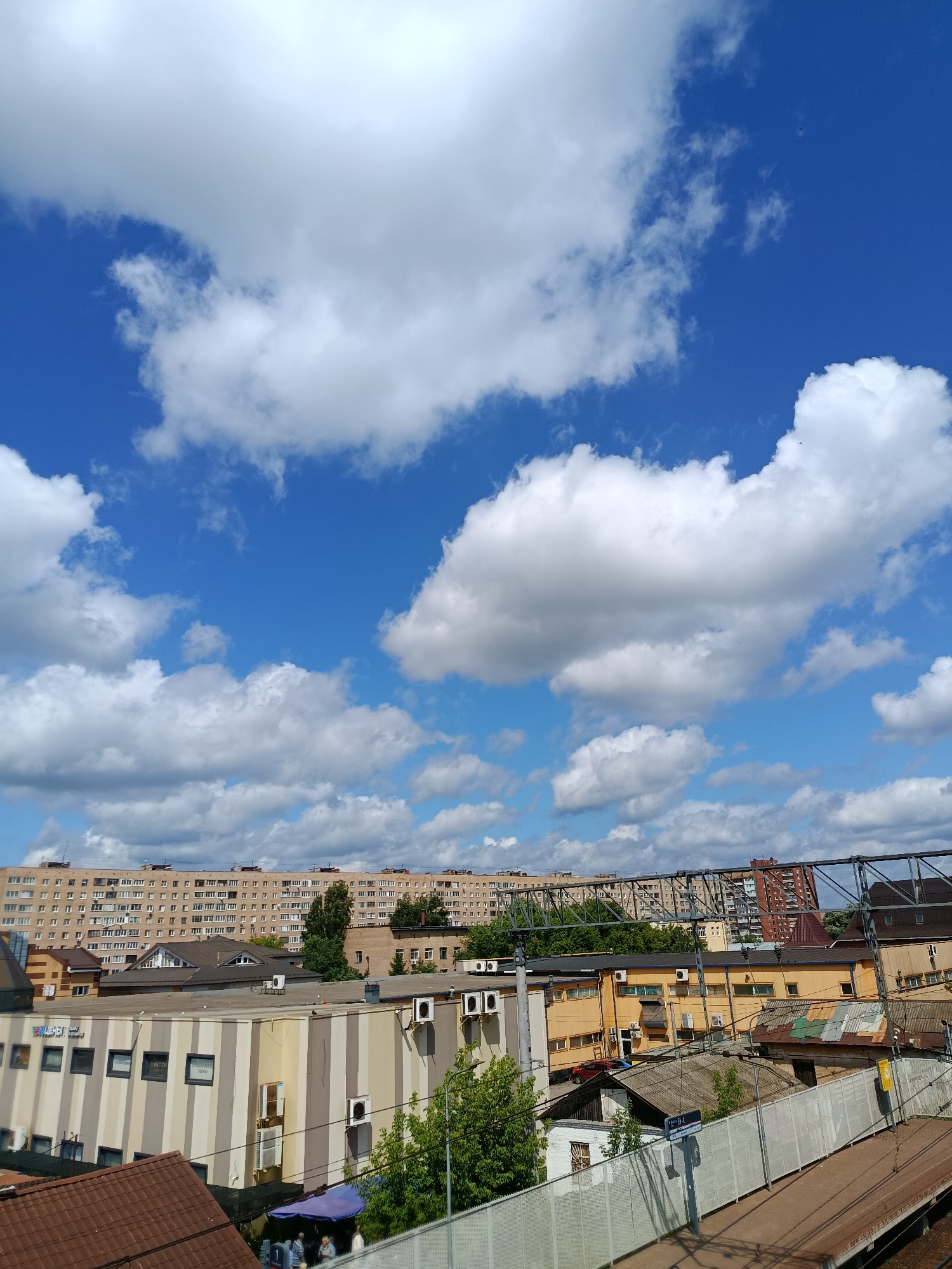 This is a medium-angle, clear, outdoor shot of an urban scene in Orekhovo-Zuyevo, Russia, under a bright blue sky dotted with fluffy white clouds. In the foreground, a white metal fence runs diagonally across the lower right, with train tracks visible beyond it. Behind the fence, there are several buildings, including a multi-story structure with a brown, shingled roof in the lower left, and a building with a striped facade of beige, yellow, and white in the center left. Further back, various industrial or commercial buildings are visible, many with yellow or beige walls and flat roofs equipped with air conditioning units. Dominating the middle ground and background are large, multi-story apartment buildings with numerous windows, typical of Soviet-era architecture. Green trees are interspersed among the buildings. Overhead, a network of dark metal structures, possibly part of an overpass or industrial framework, spans across the upper portion of the image. Two people are visible near the striped building, under a blue awning. The overall impression is one of a densely built urban environment on a sunny day.