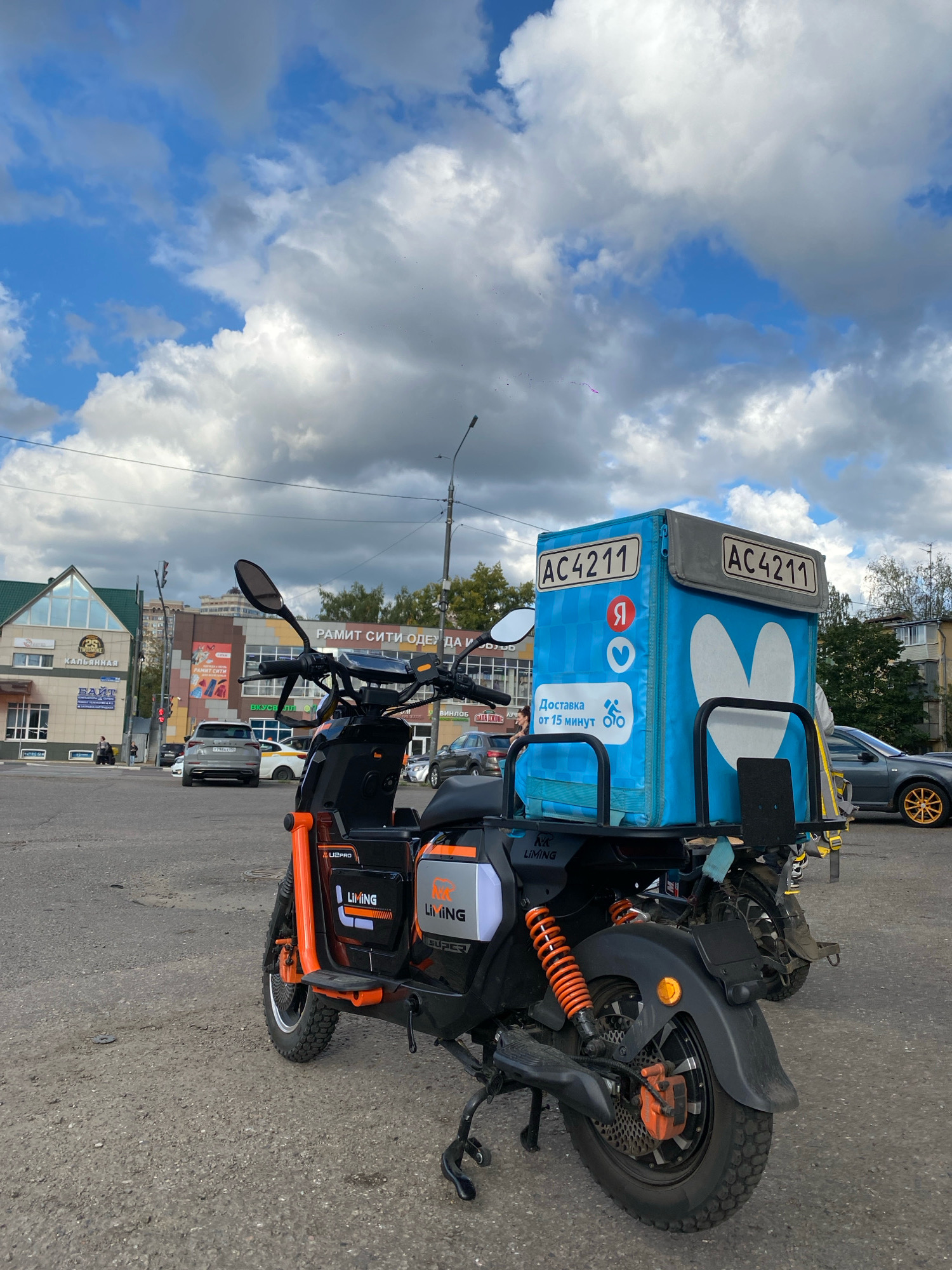 An orange and black electric scooter, a Yandex Delivery branded blue thermal bag, and two license plates with the designation AC4211 are shown in this medium shot photograph. The scooter is parked on a paved lot in front of a group of businesses. The businesses have signs with Russian text. The sky is cloudy, with patches of blue. A dark gray sedan with gold rims is parked on the right side of the image, and other cars are visible in the background. The scene is located in Ramenskoye, Russia.