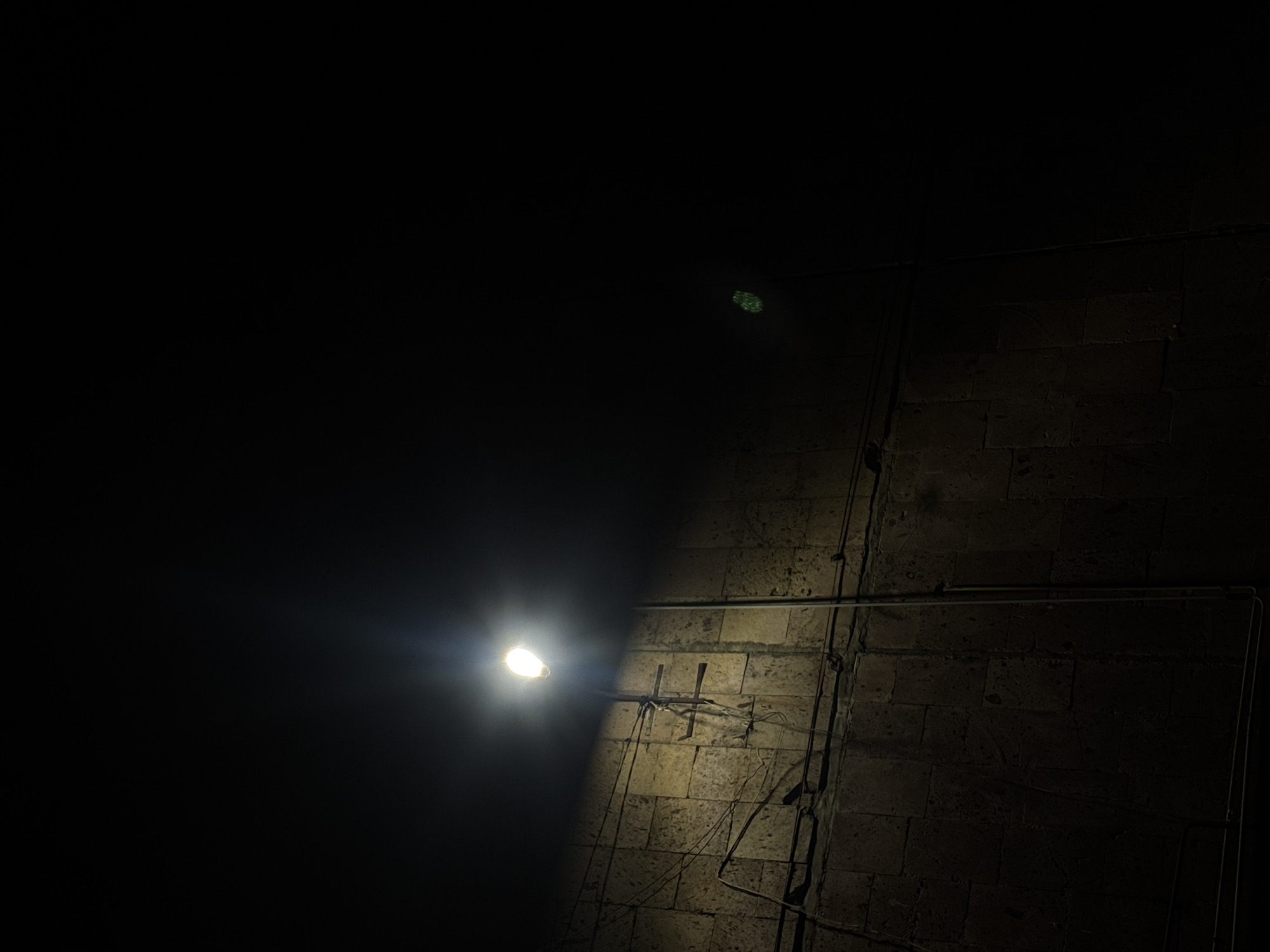 The image presents a dark outdoor scene, likely at night. The right side of the frame is dominated by a brick wall, illuminated by a nearby light source. The wall appears aged and textured. Numerous pipes and wires run along the wall's surface, adding to the scene's utilitarian and somewhat disheveled character.
To the left of the illuminated wall, the image is mostly black, indicating deep shadow or an absence of light. A bright light is visible, casting a strong glare. The surrounding area is dark, but the light suggests the presence of an artificial source, possibly a street lamp.
I cannot determine the specific location in Yerevan, Armenia, based on this image alone.