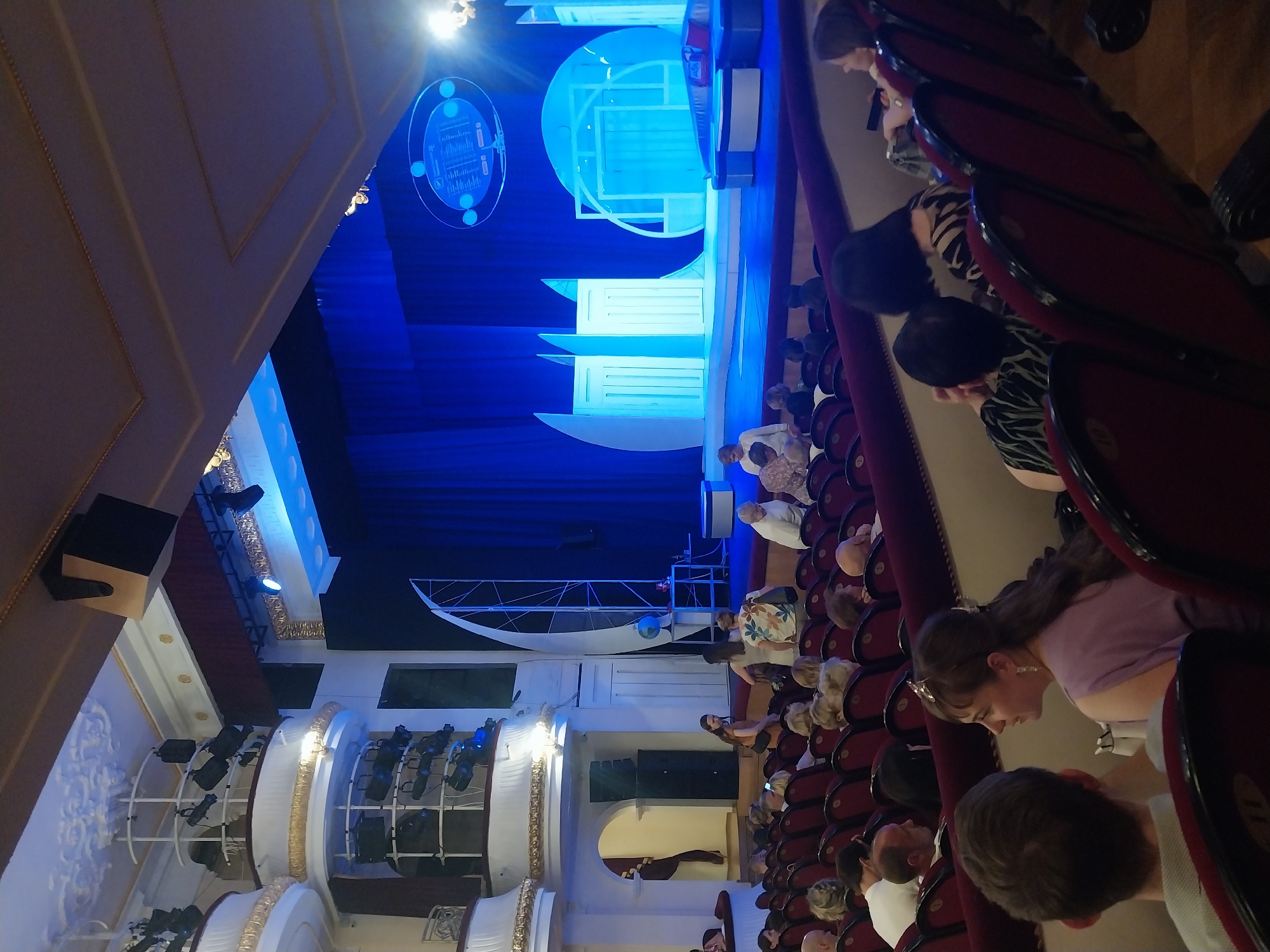 The image shows the interior of a theater in Orenburg, Russia, likely before a performance. In the foreground, rows of plush red theater seats are visible, some occupied by audience members. The audience members are diverse in age and appear to be seated and looking towards the stage. The stage itself is brightly lit with blue and white lights, featuring a modern set design with geometric shapes and blue curtains. Several spotlights and stage lighting equipment are mounted on the theater's ornate balconies. People can be seen walking on or near the stage. The architecture of the theater appears to be classic, with decorative moldings and balconies. The overall atmosphere is one of anticipation for a cultural event.