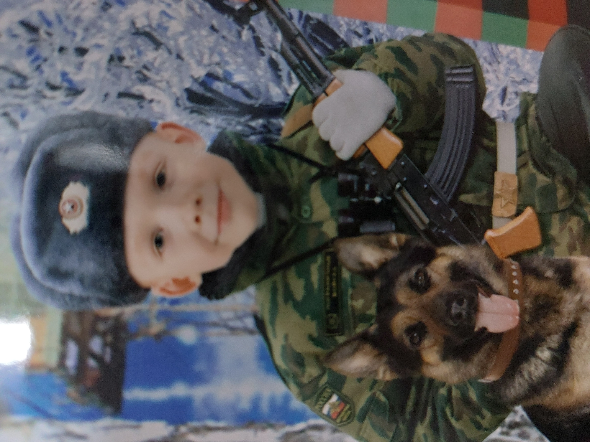 A boy in a Russian military uniform, with a traditional ushanka hat adorned with a Russian emblem, poses with a German Shepherd dog and an AK-47 assault rifle. He is wearing white gloves and a camouflage jacket. The uniform has various patches and insignias, including the Russian flag. The dog wears a studded collar and has its tongue out. The background is a blurry mix of colors and patterns, appearing to be an indoor setup, possibly a photo studio, with a backdrop of snow and a checkered flag. The image is rotated, presenting the scene sideways. The location is Chaikovsky, Russia.