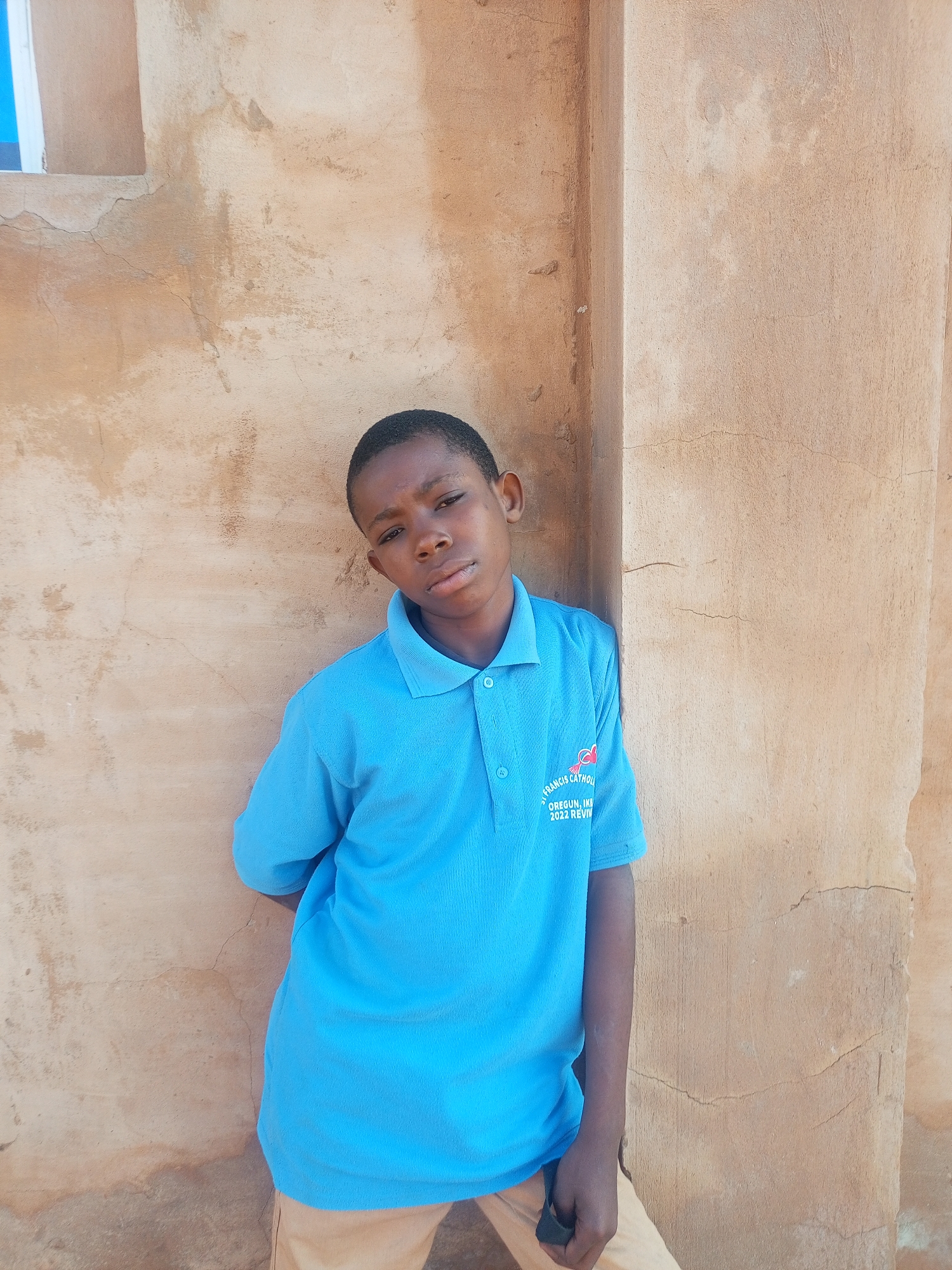 A young boy stands leaning against a textured, light brown wall. He is wearing a bright blue polo shirt with a logo on the left chest that reads "ST. FRANCIS CATHOL." and below that "OREGUN, IKEJA 2022 REVI." He also wears light brown pants. His left arm is bent and his hand is not visible. The wall has visible cracks and patches of different colors, indicating it is outdoors. The scene appears to be in a sunlit environment. The location is the exterior of St. Francis Catholic Church in Oregun, Ikeja, Lagos State, Nigeria.