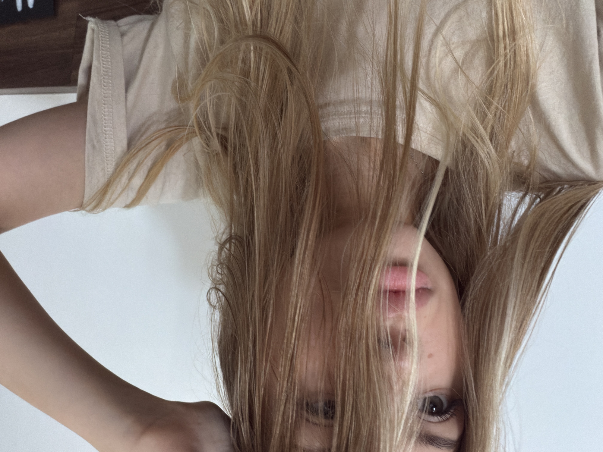 This media file features a close-up, upside-down shot of a young person with blonde hair. Their face is partially obscured by their hair, but their eye and lips are visible. They are wearing a light-colored t-shirt. The background is a plain white wall. The image appears to have been taken indoors, likely in a personal setting such as a home. There are no discernible cues for time of day, weather, or specific location within Đà Nẵng, Vietnam, beyond the general context provided. The overall mood is casual and intimate.