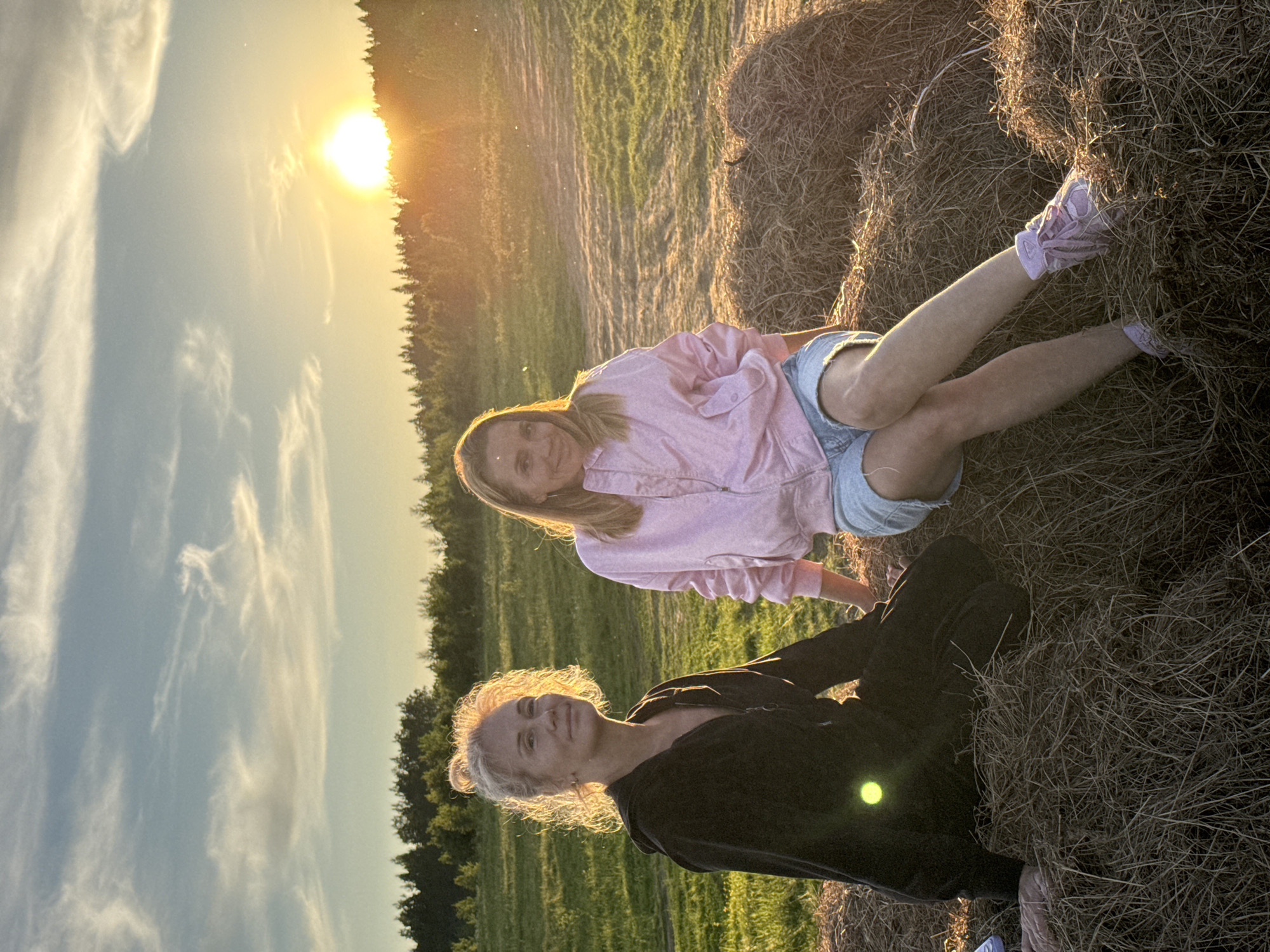 This image depicts two women in a rural field during the golden hour of sunset. The bright sun, positioned low on the left side of the frame, casts a warm, golden light across the scene, creating a strong backlight that illuminates the subjects and causes a lens flare. The sky above is a clear blue adorned with wispy white clouds.
In the foreground, a woman with light, curly hair and a dark (possibly black) long-sleeved top is seated, looking directly at the camera with a relaxed, pleasant expression. Her hair is brightly backlit by the setting sun, giving it a luminous halo effect. Her light pink shoes are partially visible. Behind her and slightly to the right, another woman with long, straight blonde hair is reclining on a large, round hay bale. She is wearing a pink long-sleeved top and light blue denim shorts, with light pink sneakers visible on her feet. She is also looking towards the camera with a gentle smile.
The setting is an open field with green grass, leading towards a line of dark trees that form the horizon. The large hay bale occupies a significant portion of the right side of the frame, adding to the agricultural character of the scene. The overall atmosphere conveyed is peaceful and relaxed, capturing a moment of enjoyment in nature. There is no visible text in the image. This scene is located in a field near Стеклянный, Russia.