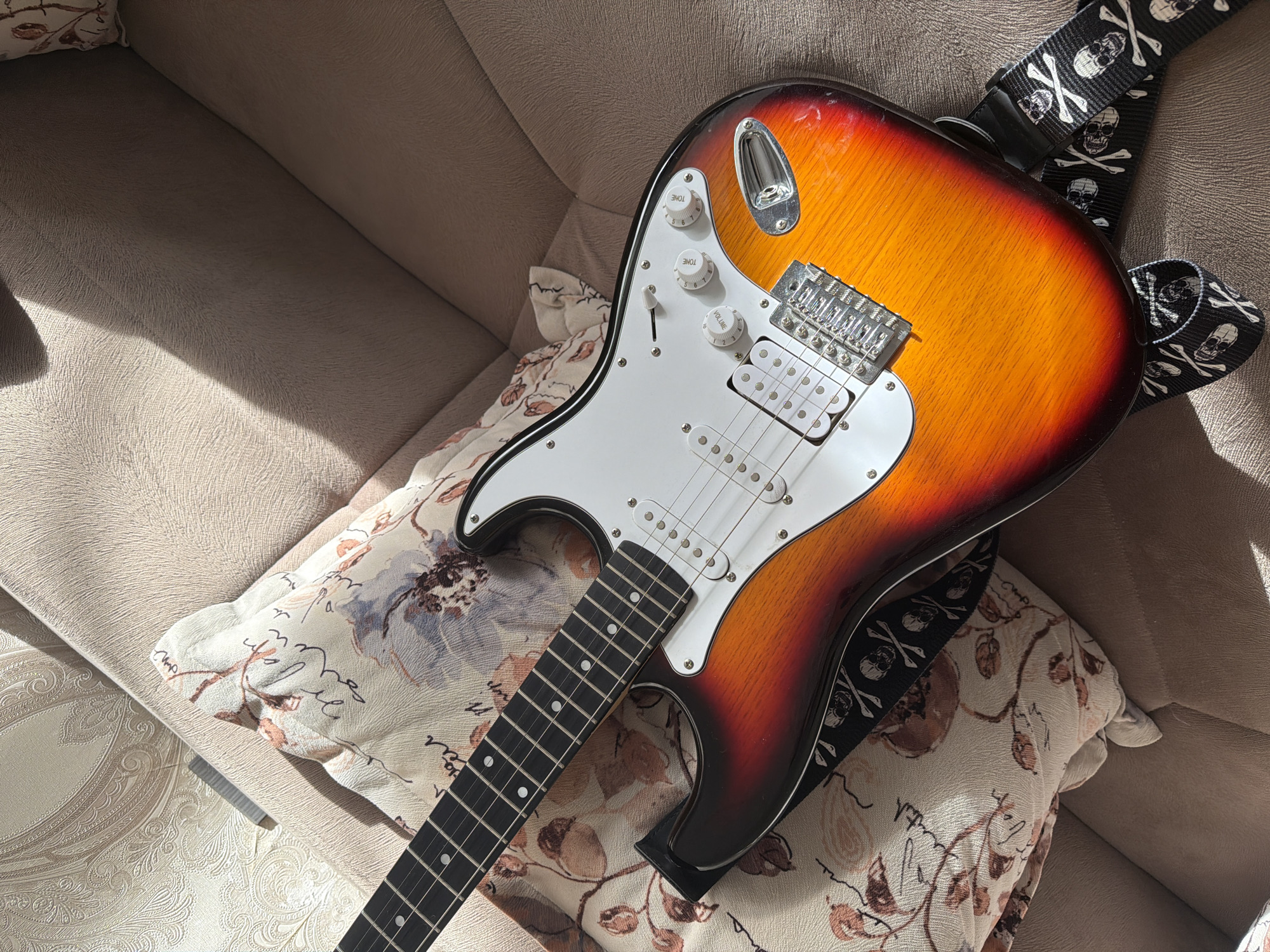 An electric guitar with a sunburst finish rests on a light-colored couch. The guitar has a white pickguard and a black strap with a skull and crossbones pattern. The couch is covered in a textured fabric, and a pillow with a floral and script pattern is visible. The scene is lit by natural light, casting shadows across the couch and guitar. The image appears to be taken indoors. No people are present in the image. The location is not specified to be in Baku City, Azerbaijan.
