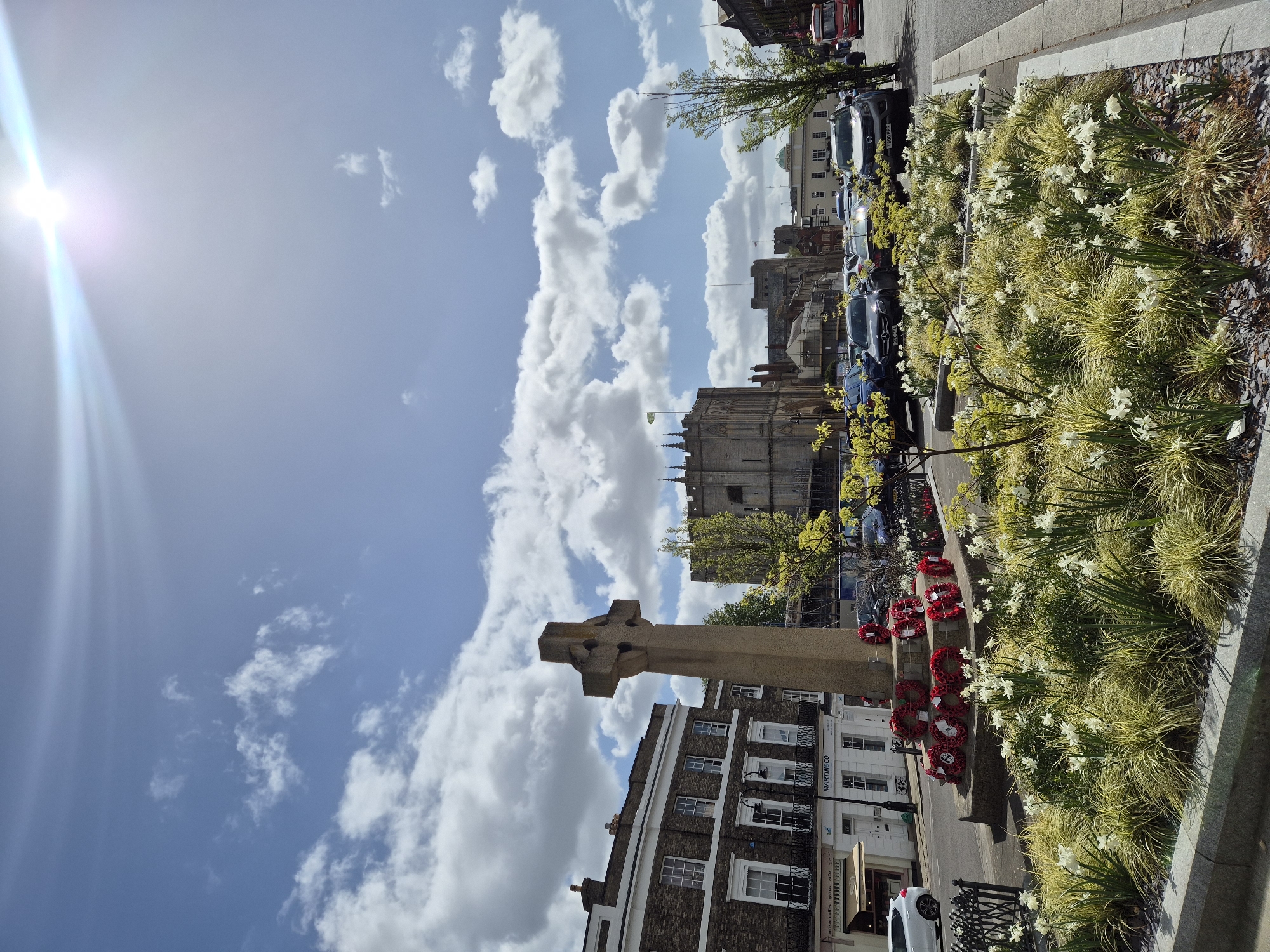 This photo depicts a street scene with a Celtic cross war memorial in the foreground, likely in a town within West Suffolk, UK. The memorial is adorned with several red poppy wreaths. Behind the memorial is a street with buildings, parked cars, flower beds, and hints of taller structures in the distance. The sky is blue with cumulus clouds and the sun is visible, casting bright light. The image is oriented vertically.