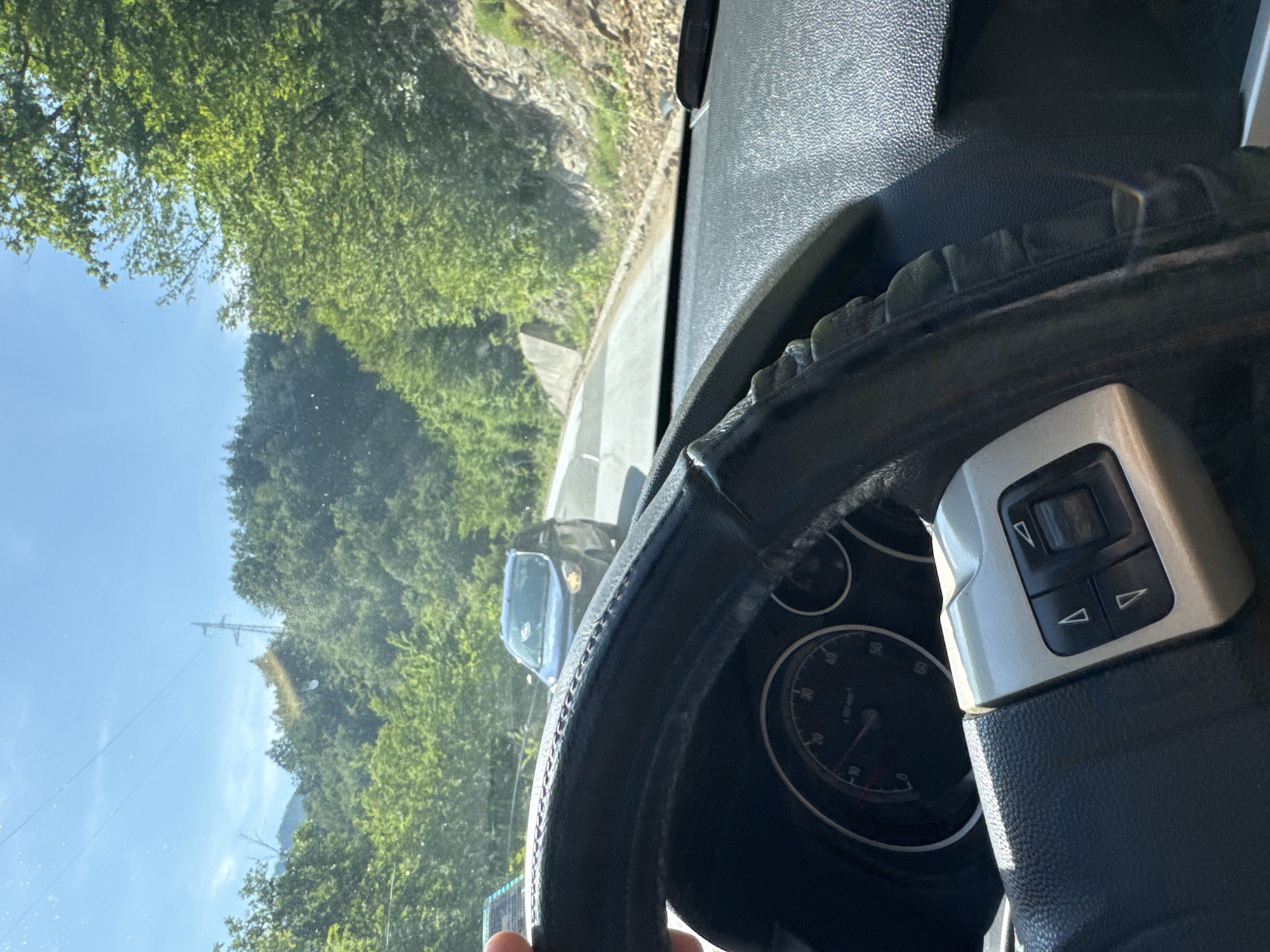 The image presents a first-person view from inside a car, looking out through the windshield onto a mountainous, natural landscape. The scene is set on a clear, bright, and sunny day, likely during daylight hours.
In the foreground, a black steering wheel with a textured cover occupies the lower and right portions of the frame. A hand lightly grips the bottom left of the steering wheel. On the right side of the steering wheel, a silver control module features black buttons with white arrow icons. Partially visible behind the steering wheel is a portion of the car's instrument cluster, showing what appears to be a speedometer or tachometer. The black dashboard extends across the mid-right of the image.
Through the windshield, a lush, green environment dominates the background. Dense foliage and trees cover rolling hills or mountains, suggesting a journey through a rural or natural area. A grey concrete road barrier runs alongside the road. Ahead and slightly to the left, another dark-colored car, possibly blue or black, is visible, either parked or traveling in the opposite direction. In the upper part of the sky, a utility pole with power lines is visible, partially obscured by tree branches. Some reflections or specks of dust are noticeable on the windshield. The overall impression is of driving through a verdant, hilly region.
No discernible text is clearly visible on any objects within the frame.