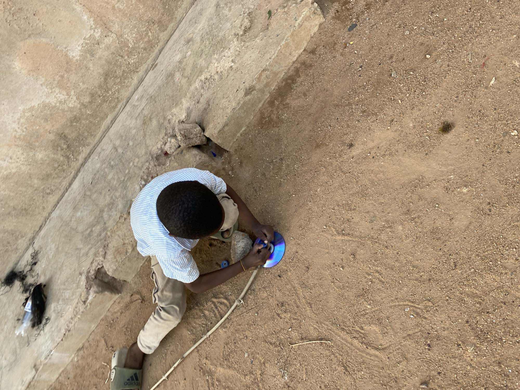 The image is taken from an overhead perspective, showing a young boy playing with a CD on a dirt surface in Bukuru, Nigeria. The boy has dark hair and skin, wearing a blue and white striped shirt and khaki pants. He is on the ground, holding the CD in his hands. Next to him are some debris, a stick, and a pair of green Adidas sandals on his feet. The ground surface is a mix of dirt and small pebbles. In the background, there's a concrete structure. To the left of the boy is some discarded plastic packaging and what appear to be hair extensions.