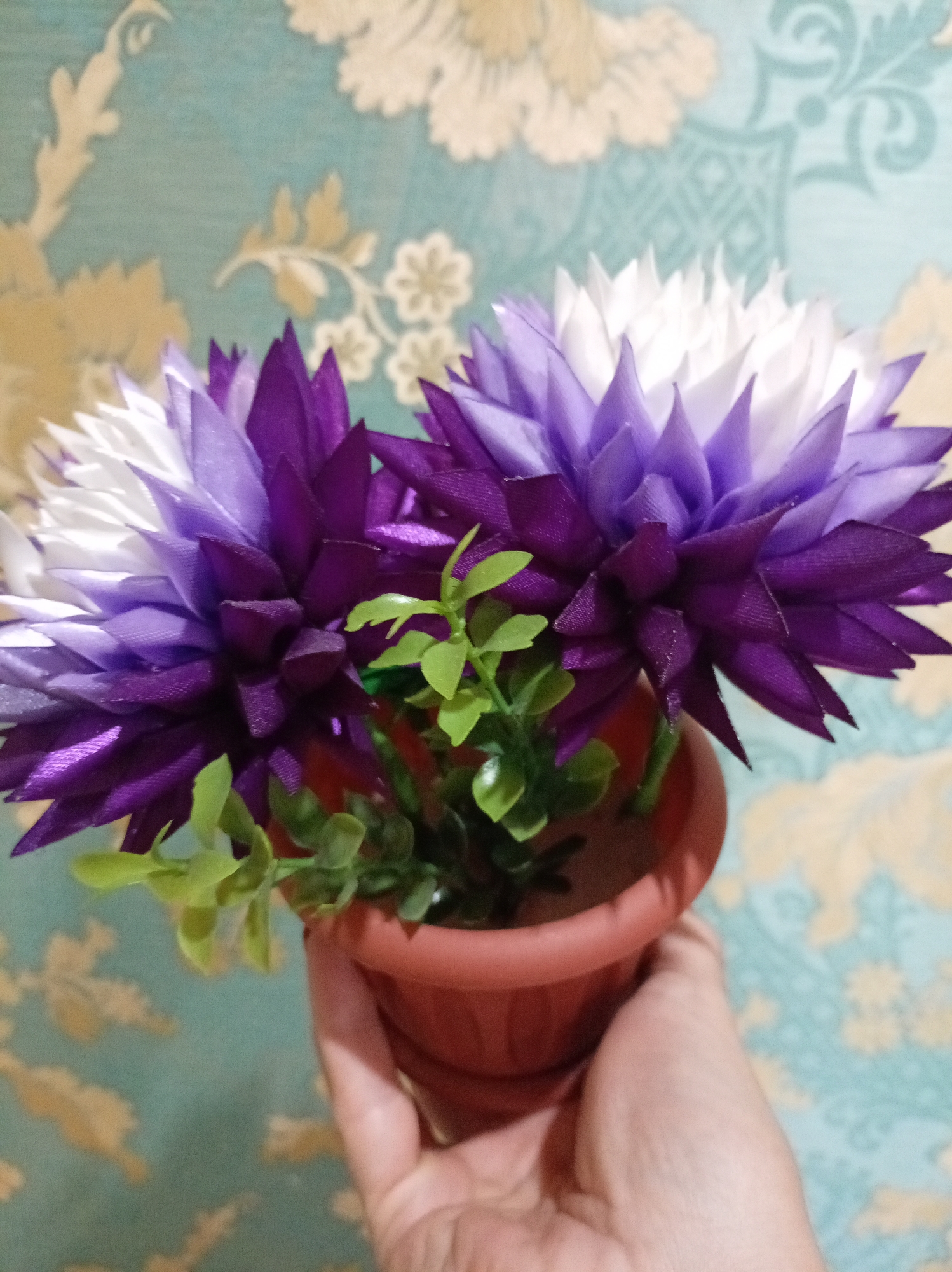 A hand holds a small terracotta pot containing two artificial flowers and green plastic leaves. The flowers have layered petals, transitioning from deep purple at the base to lighter shades of purple and then white at the tips. The background is a wall covered in wallpaper with a repeating pattern of stylized floral motifs in muted cream and blue-green tones. The lighting appears to be indoor, with no indication of time of day or weather. The scene is static, with no discernible activity taking place beyond the presentation of the potted flowers. There is no visible text in the image. The location is identified as Bologoe, Russia.
