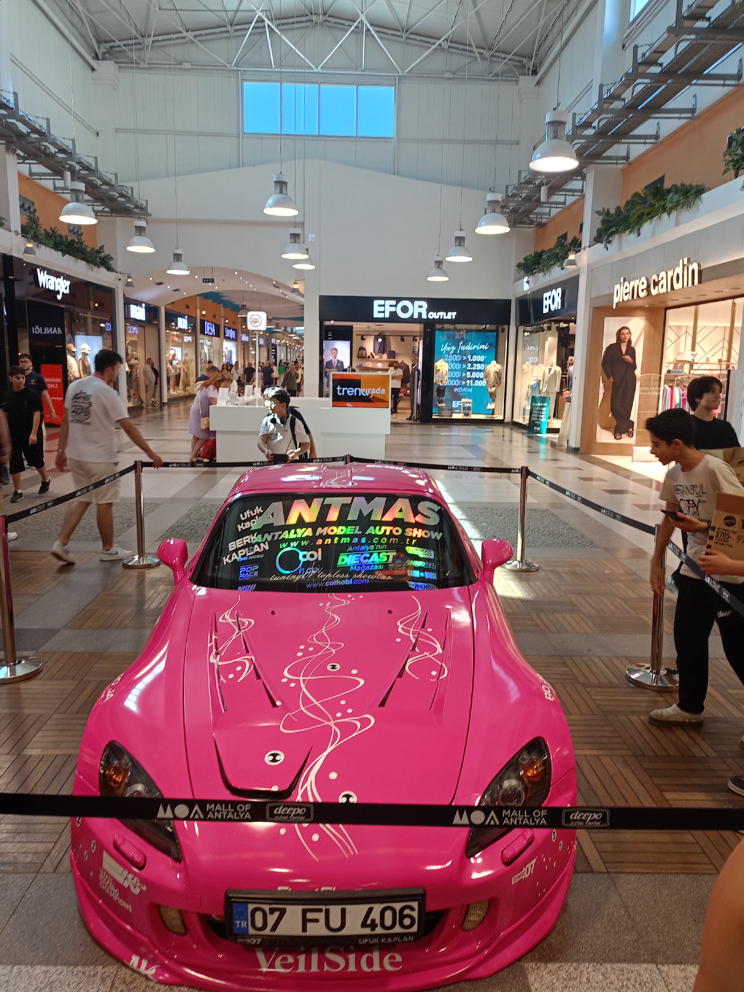 The image shows a bright pink sports car, possibly a Honda S2000, parked indoors at the Mall of Antalya, located in Kepez, Turkey. The car is decorated with white swirling decals and has a custom license plate that reads "07 FU 406". There are several people in the background, some browsing in the shops and others standing near the car. The mall interior features a high ceiling with skylights and multiple storefronts. A black rope barrier is set up around the car, indicating it is part of an exhibit or event. The overall scene suggests a public event or display within a shopping mall.