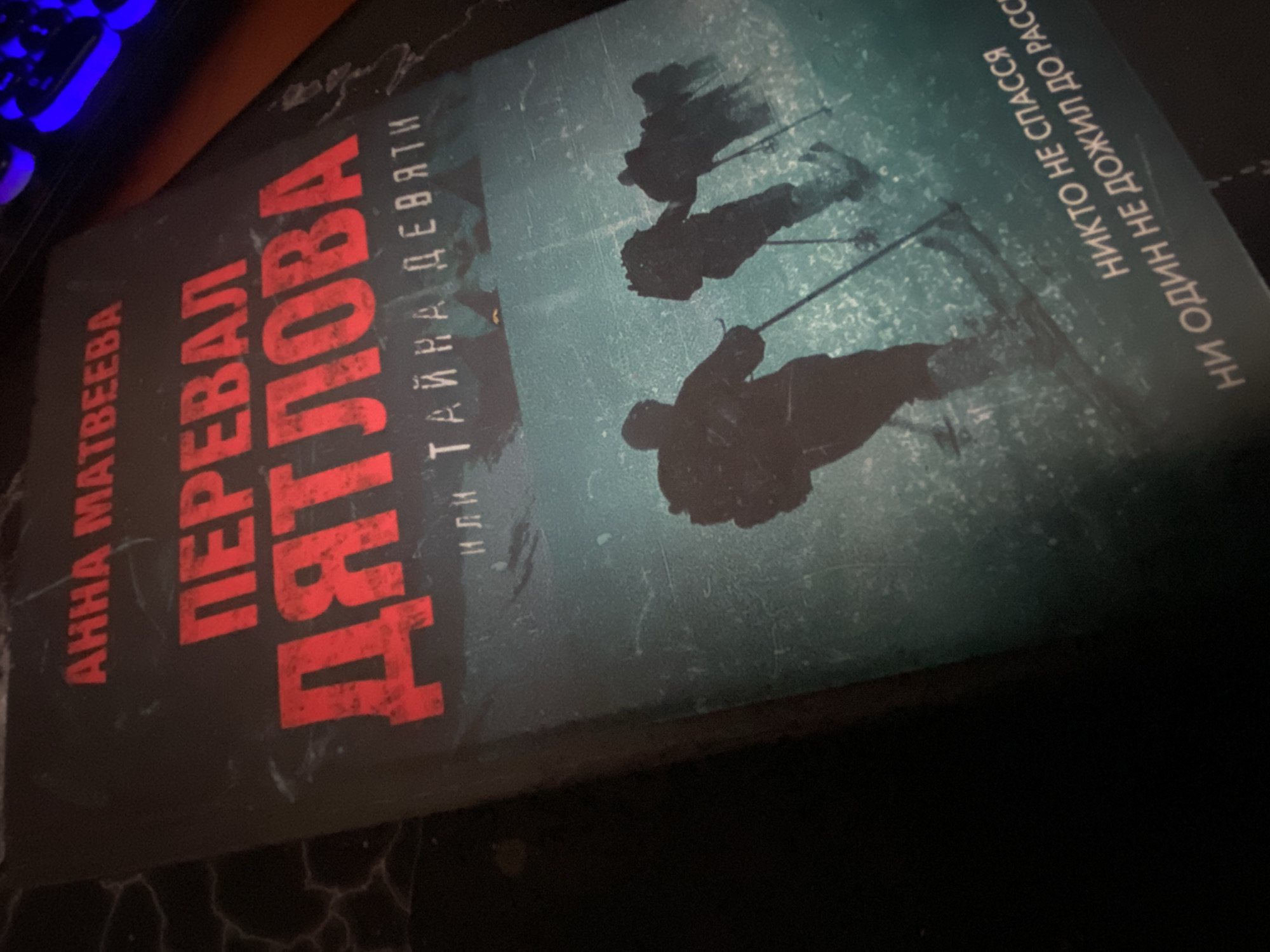 The image shows a book titled "Перевал Дятлова" (Dyatlov Pass) by Anna Matveeva. The cover is predominantly dark with red lettering for the title. There are silhouette-like figures of people skiing on a teal-colored background. The book is laying on a dark surface. A portion of a blue, illuminated keyboard is visible in the upper left corner. There is no mention of Парэчча, Belarus.