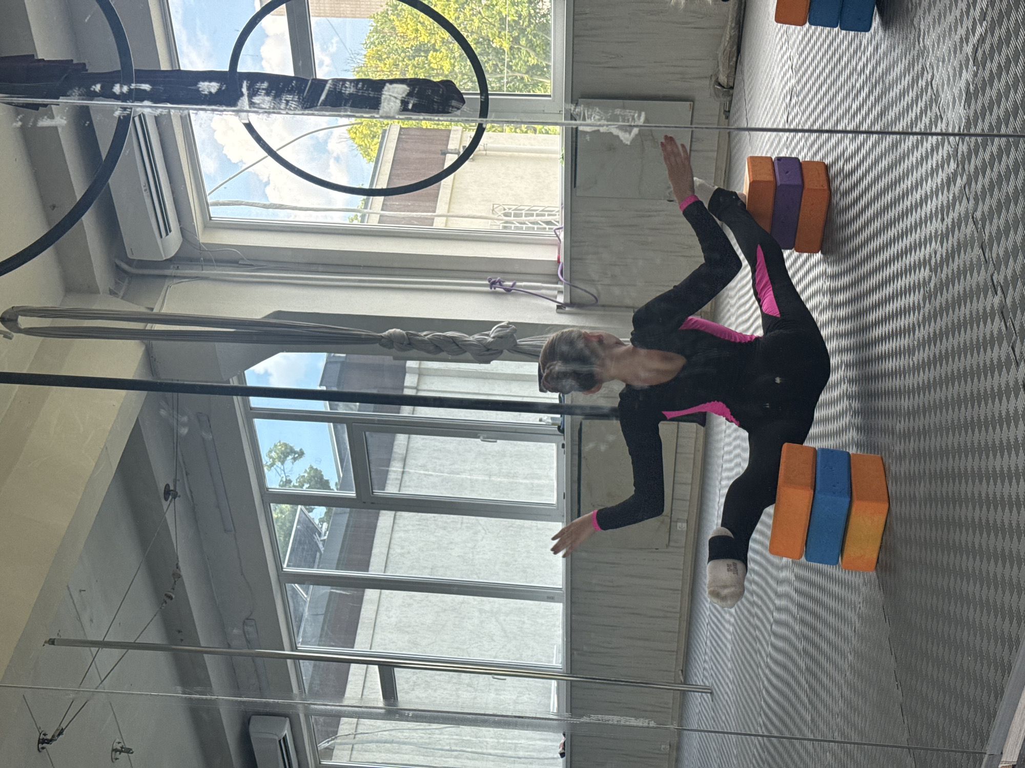 The image depicts a woman in a black and pink leotard performing a split on stacked yoga blocks in an indoor studio. The studio has large windows offering a view of trees and a blue sky. The floor is covered with a grey puzzle mat. A pole and aerial silks are visible in the background, suggesting the space is used for pole dancing or aerial acrobatics. The scene is reflected in a large mirror, which occupies the left side of the frame. The overall impression is one of a fitness or dance studio. The location of Dnipro, Ukraine is not evident from the image itself.