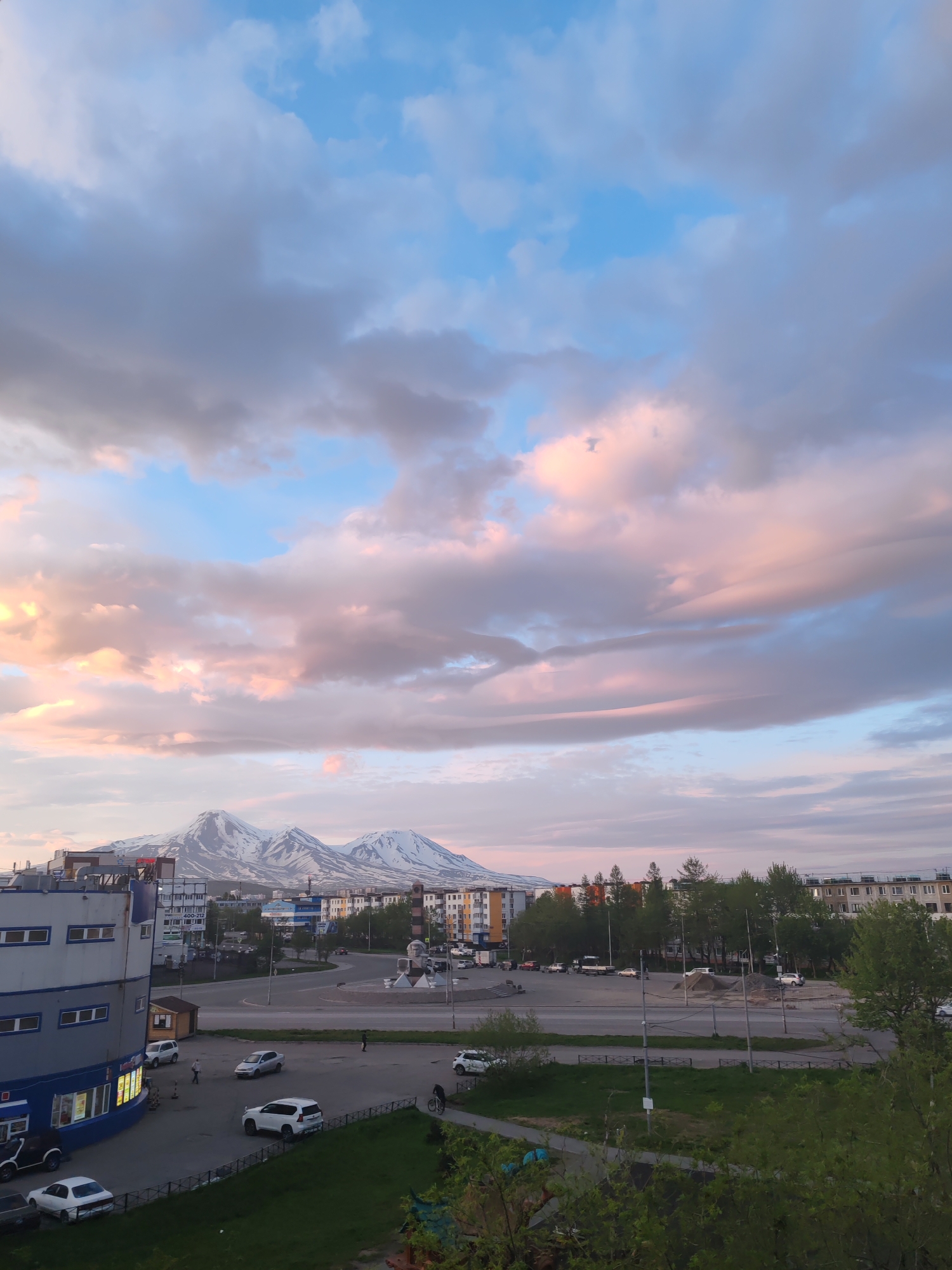 The image depicts a cityscape with snow-capped mountains in the background, characteristic of Petropavlovsk-Kamchatsky, Russia. In the foreground, a multi-story building with blue and white siding is visible. Several cars are parked in a lot next to the building, and a few people are walking. The street in front of the building shows light traffic, with vehicles driving and parked. Beyond the street, more apartment buildings and trees line the horizon. The sky above is filled with dramatic clouds, tinged with pink and orange hues, suggesting either sunrise or sunset. The overall scene conveys a sense of urban life set against a dramatic natural backdrop.