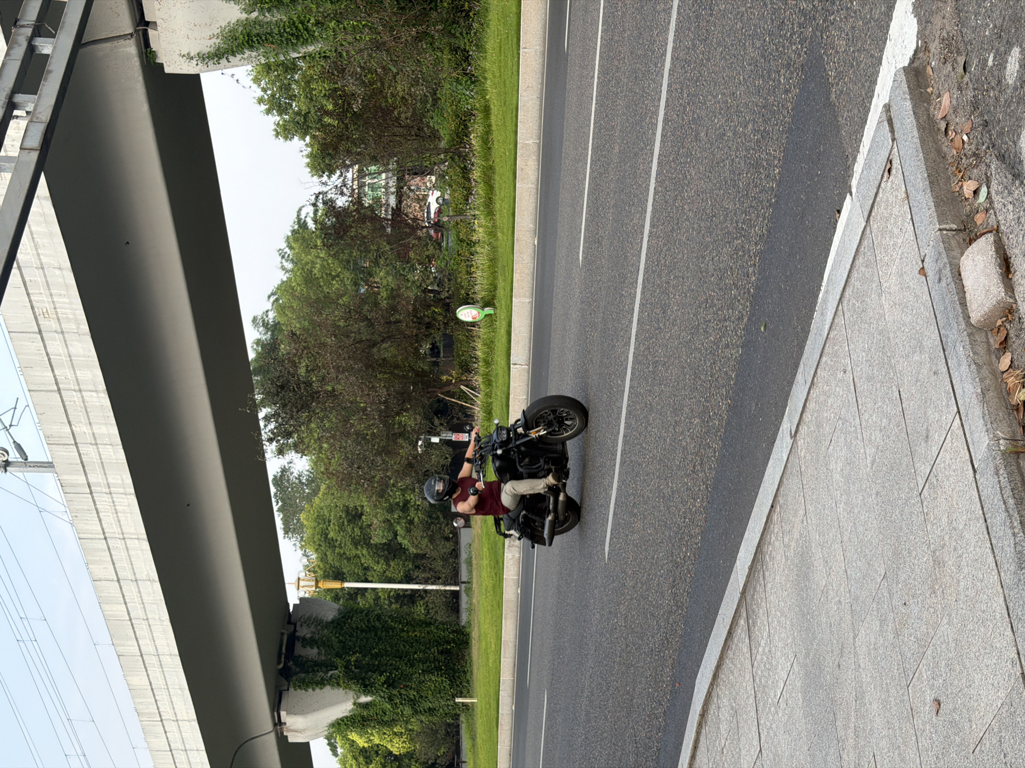 A lone motorcyclist is riding a black cruiser motorcycle down a multi-lane asphalt road. The rider is wearing a maroon tank top, light-colored pants, and a black helmet. They are positioned in the right lane, facing towards the right side of the frame.
The road is bordered by a sidewalk made of large, light-colored stone pavers on the left. The right side of the road features a grassy median with several trees and some dense green foliage. A concrete overpass, likely part of an elevated roadway or railway, spans across the upper portion of the image, supported by large concrete pillars. One pillar on the left is heavily covered in green vines. A bright yellow lamp post with decorative accents is visible between the overpass and the trees on the left. The sky is overcast and pale blue. In the background, beyond the trees, a multi-story building with green accents and a white facade is partially visible. There are also cars parked or driving in the distance. The scene appears to be in an urban or suburban environment in Jiangjin, China.