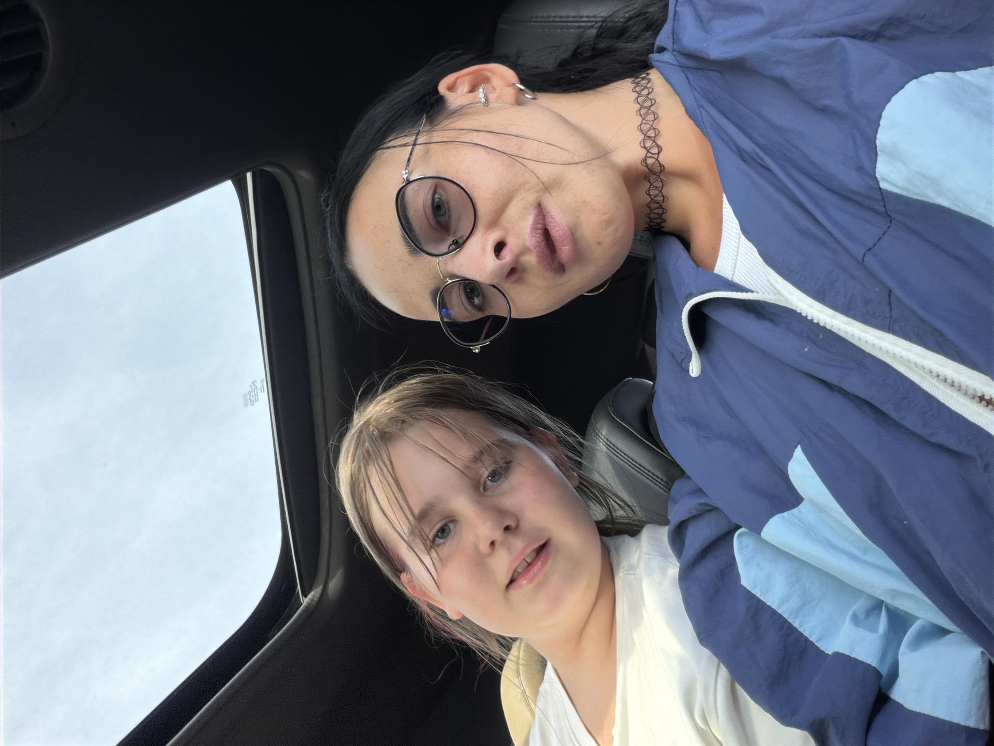 Two individuals are inside a vehicle, with the car's sunroof open above them. The person in the foreground, a woman with dark hair, wears round glasses, a black choker necklace, and a blue and light blue jacket over a white top. To her left is a younger individual, likely a girl, with light brown hair and blue eyes, wearing a white t-shirt. The background shows the interior of the car, including black car seats and a glimpse of the sky through the sunroof. The scene is set in a public place in the city of Sholokovo, Russia.