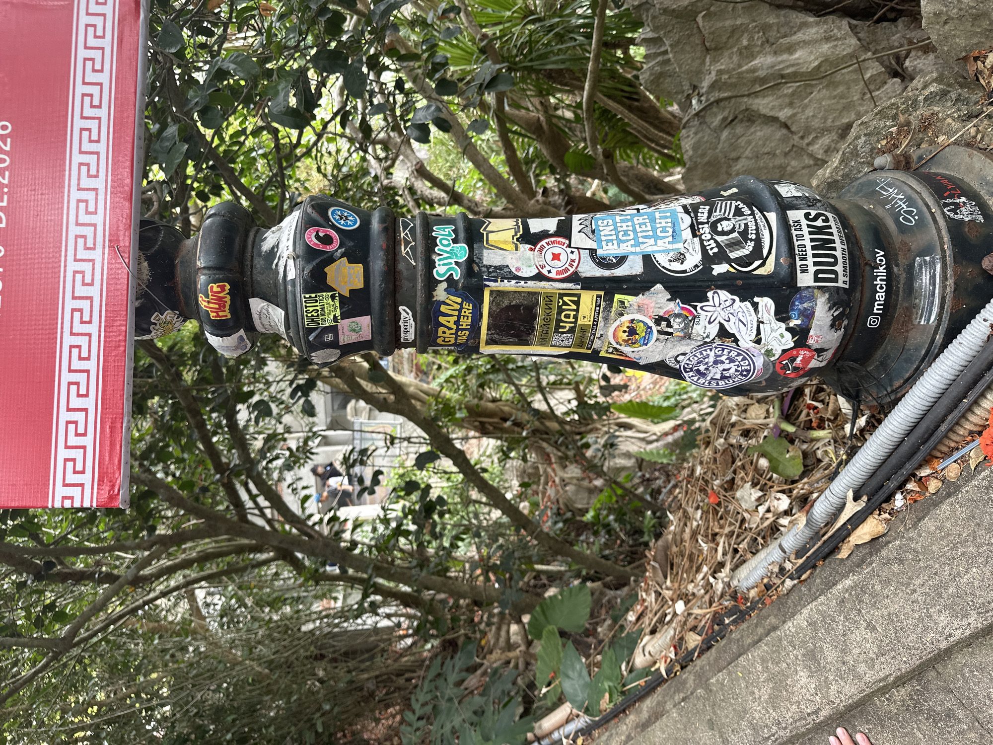 A black lamppost is covered in stickers of various sizes and designs. The lamppost is located outdoors, next to a concrete walkway and a retaining wall. Behind the lamppost, there is dense foliage and trees. A red banner with a white geometric pattern is visible at the top of the image. The stickers include text in English, Russian, and German, as well as images of logos, symbols, and faces. The environment suggests an urban or semi-urban setting. The image does not provide specific information to confirm a location in Đà Nẵng, Vietnam. There are no people clearly depicted in the image.