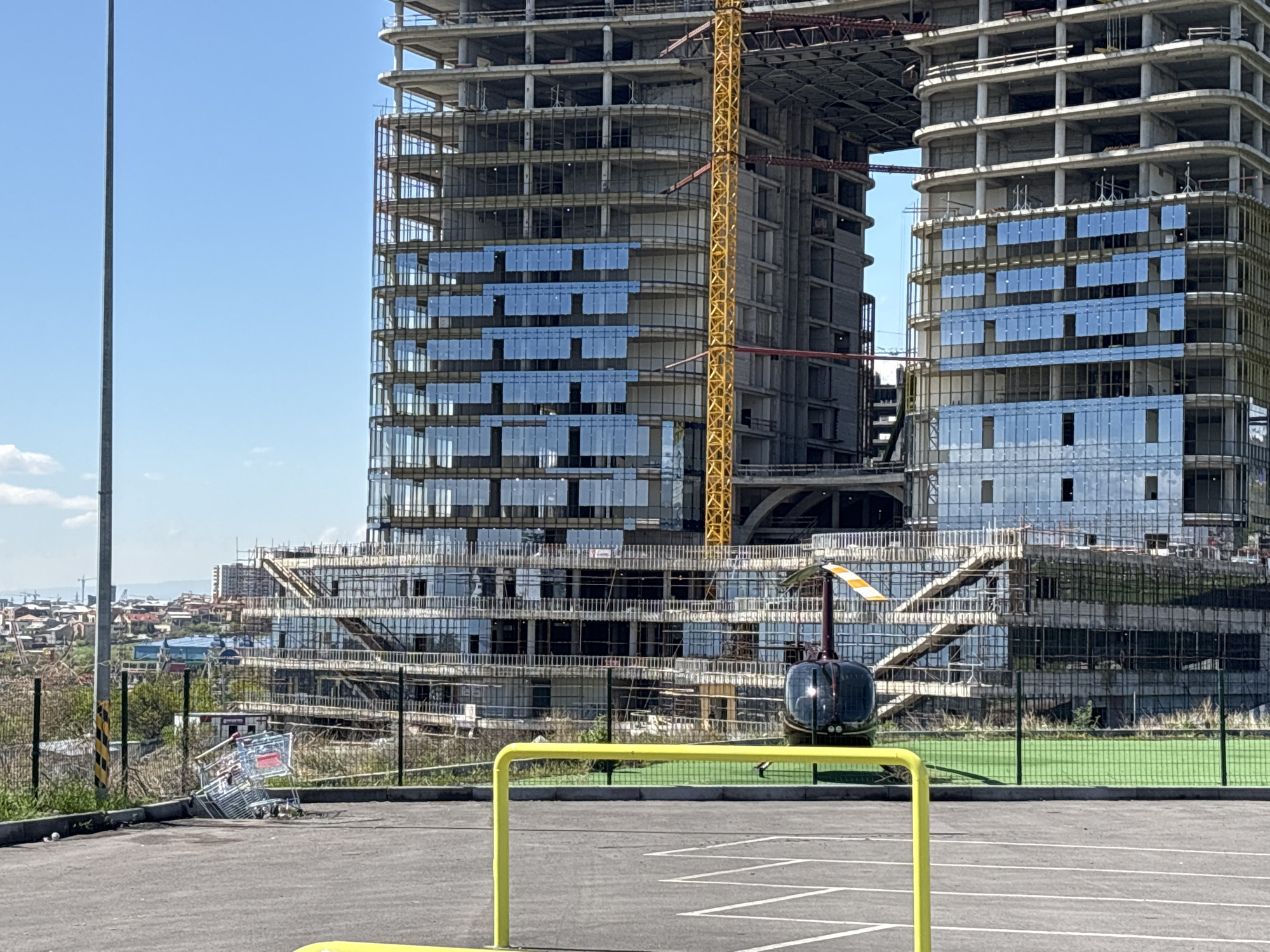 The image shows a partially completed construction project of two tall buildings connected by a bridge, located in Yerevan, Armenia. The buildings appear to be in the process of having glass panels installed. A yellow crane is visible between the two buildings. In the foreground, a black helicopter is parked on a paved surface with yellow railings. Beyond the paved area, there is a green space and a fence. To the left, there is a slope with vegetation and an overturned shopping cart. In the distance, there is a view of the city of Yerevan under a clear blue sky.