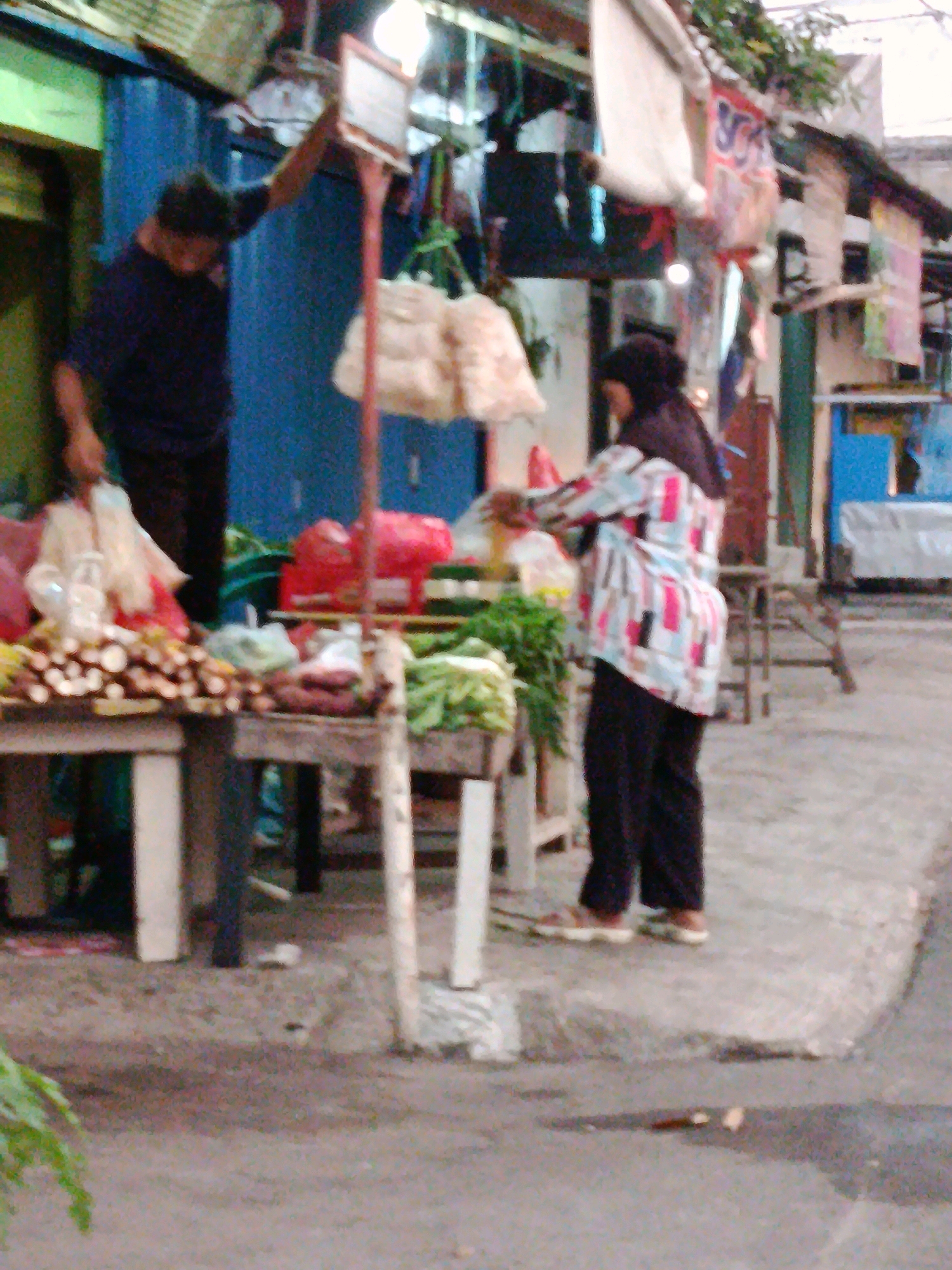 In Bekasi, Indonesia, a blurry, medium-shot image captures a street-side market scene. A person wearing a dark shirt leans against a blue stall, their face obscured. In front of them, a table displays a pile of sliced, light-colored root vegetables, possibly ginger or turmeric, alongside plastic bags containing food items. To the right, another person, wearing a headscarf and a colorful, striped shirt over dark pants, stands near a table laden with green leafy vegetables and other produce. The background reveals more market stalls with hanging fabrics and merchandise, suggesting a bustling, local commercial area. The overall impression is that of a typical day at a roadside market in a densely populated urban setting.