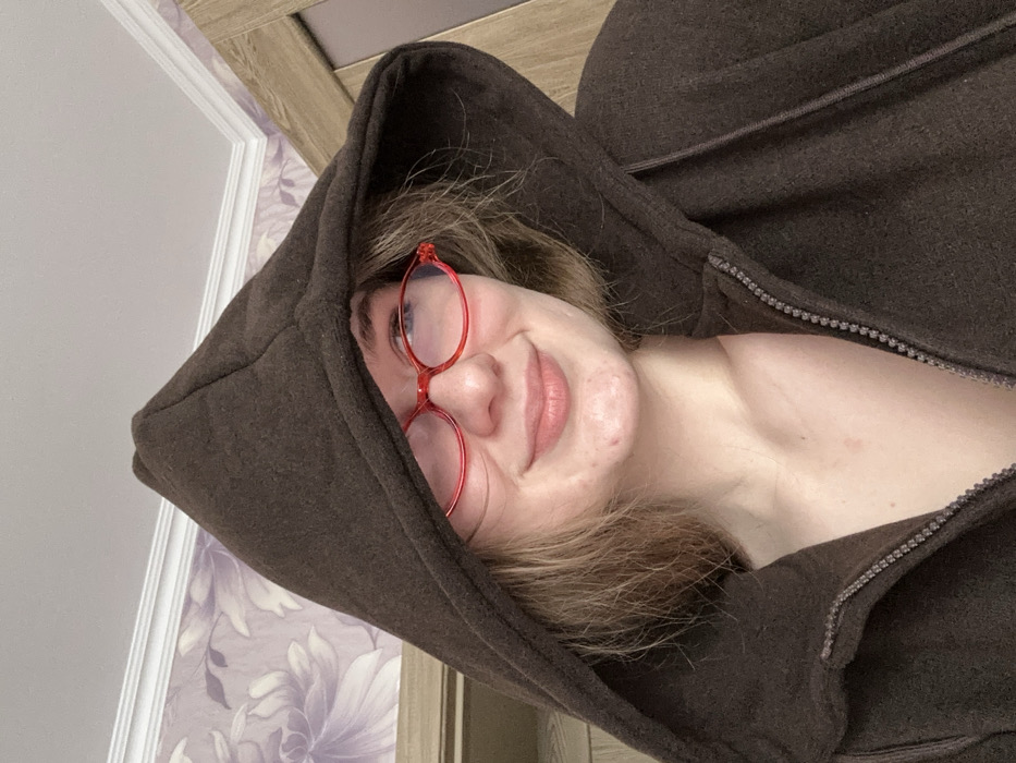 A close-up, head-and-shoulders image features an individual, likely female, within an indoor residential setting, reported to be located in Nizhnekamsk, Russia. The person wears a dark brown, possibly fleece, hooded sweatshirt with the hood pulled over the head, partially obscuring light brown hair. Red, oval-framed eyeglasses are worn, and a slight, closed-lip smile is directed slightly to the viewer's right. The sweatshirt is partially unzipped, revealing a portion of the upper chest. Minor skin blemishes are visible on the chin and cheek. The background consists of a wall with light-toned floral wallpaper featuring larger, darker floral patterns on the left, a white crown molding along the ceiling line, and a segment of a light-colored wooden door frame or door on the right.