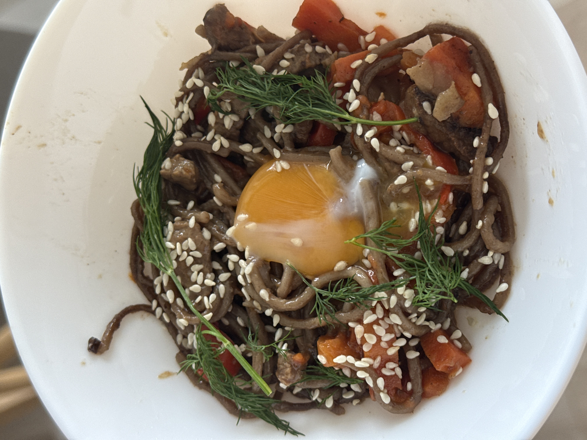A high-angle shot depicts a white bowl containing a dish of soba noodles. The noodles are dark in color and tossed with diced carrots and what appears to be sauteed mushrooms. A raw egg yolk sits at the center of the noodles, slightly glistening. Sprigs of fresh dill are artfully arranged around the edge of the dish. Sesame seeds are liberally sprinkled across the top of the noodles. The bowl is situated on a surface with muted colors. No specific location or individuals are visible in the image.