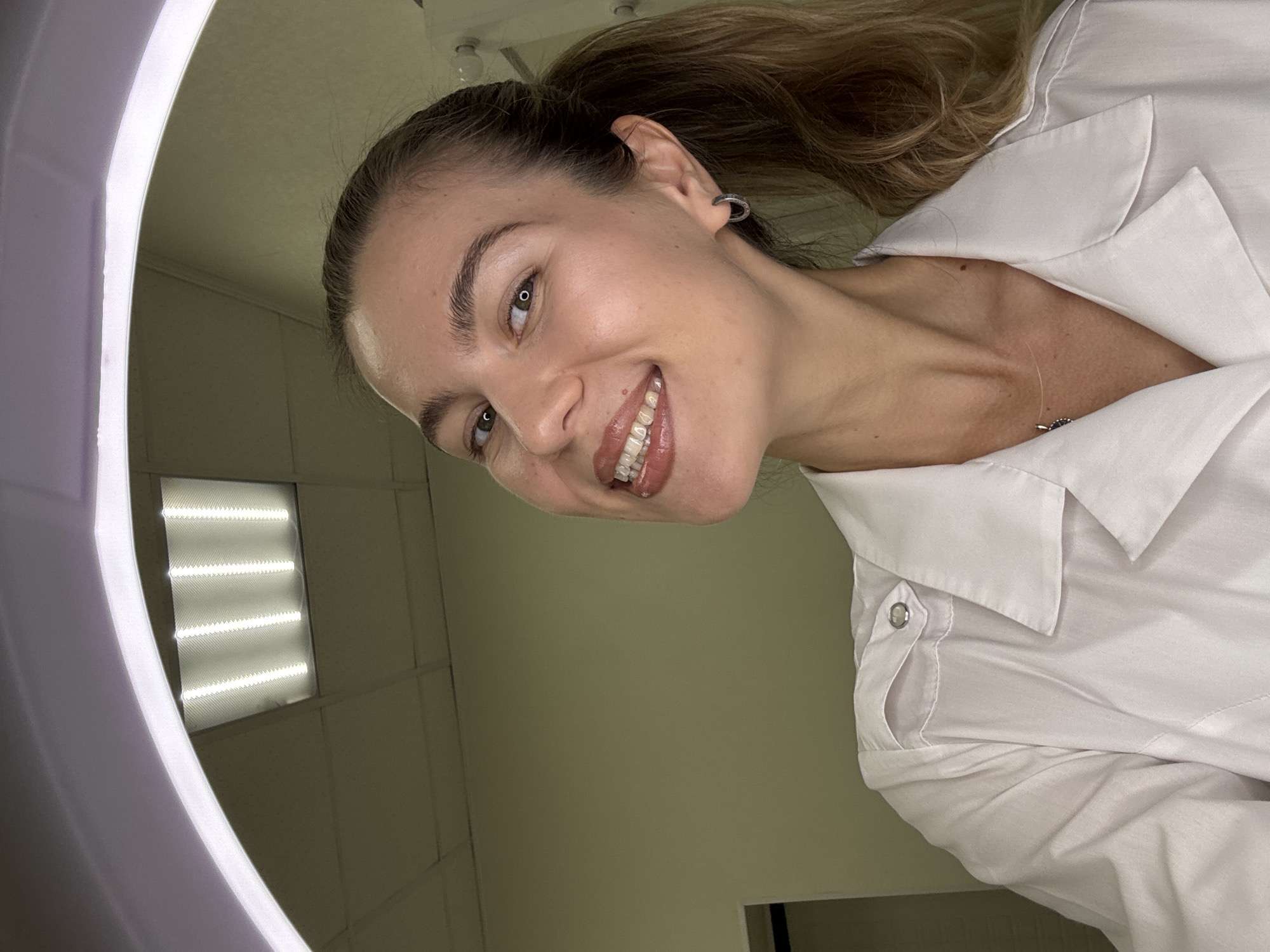A woman with blonde hair tied in a ponytail and a light-colored collared shirt is shown from the chest up. She is smiling broadly, revealing clear braces on her teeth. The woman is wearing a small hoop earring in her left ear and a delicate necklace with a small pendant. The background is a plain wall and a ceiling with fluorescent lighting. A ring light is visible around the top of the frame.
