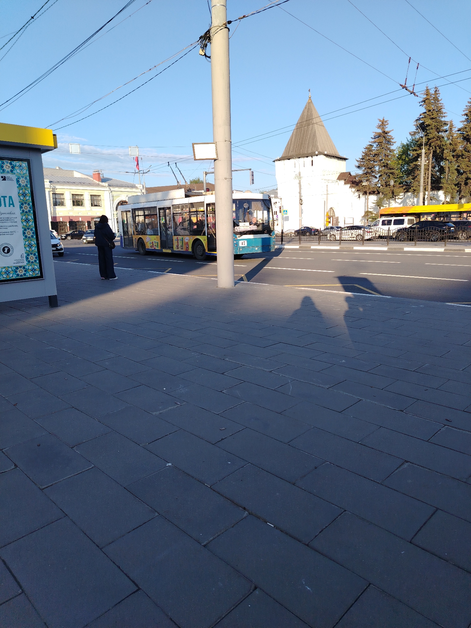 The image captures a street scene in Rusanovo, Russia. A person dressed in black stands on a paved sidewalk next to a bus stop. A yellow and blue bus is paused in the road, with people visible through the windows. Behind the bus, there is traffic with various vehicles. In the background, a white building with a pyramid-shaped roof stands out, along with evergreen trees. Overhead power lines are strung across the sky, which appears clear and blue. The light casts shadows of the person and other objects onto the sidewalk.