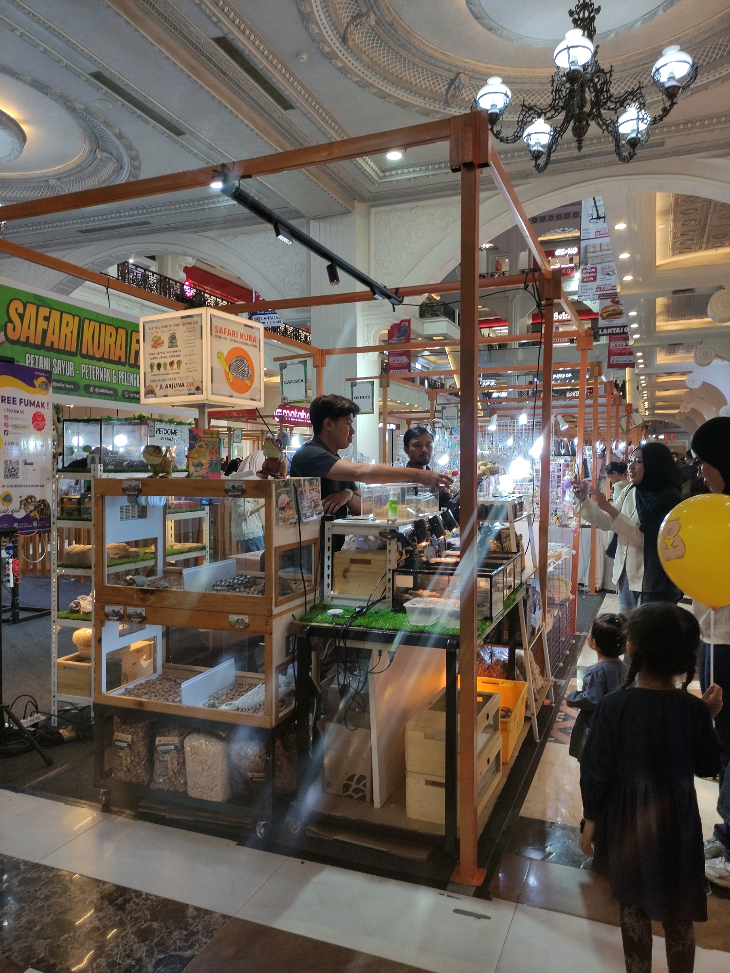 The image depicts an indoor market stall, possibly in Sinduadi, Indonesia, specializing in reptiles and related supplies. In the foreground, two young girls, one in a dark blue dress and the other with braided hair, are looking towards the stall. A woman in a black hijab and a cream jacket stands to their right, holding a yellow balloon with a cartoon bear illustration. To the left of the stall, a man in a dark shirt is serving customers.
The stall itself is constructed with wooden frames and features multiple shelves displaying terrariums with small animals, likely reptiles, and bags of substrate or food. A large green banner with the text "SAFARI KURA" is visible on the left. The background reveals more market stalls and architectural elements of what appears to be a shopping mall or indoor complex, including decorative ceilings and chandeliers. The lighting is bright, suggesting an indoor commercial setting.