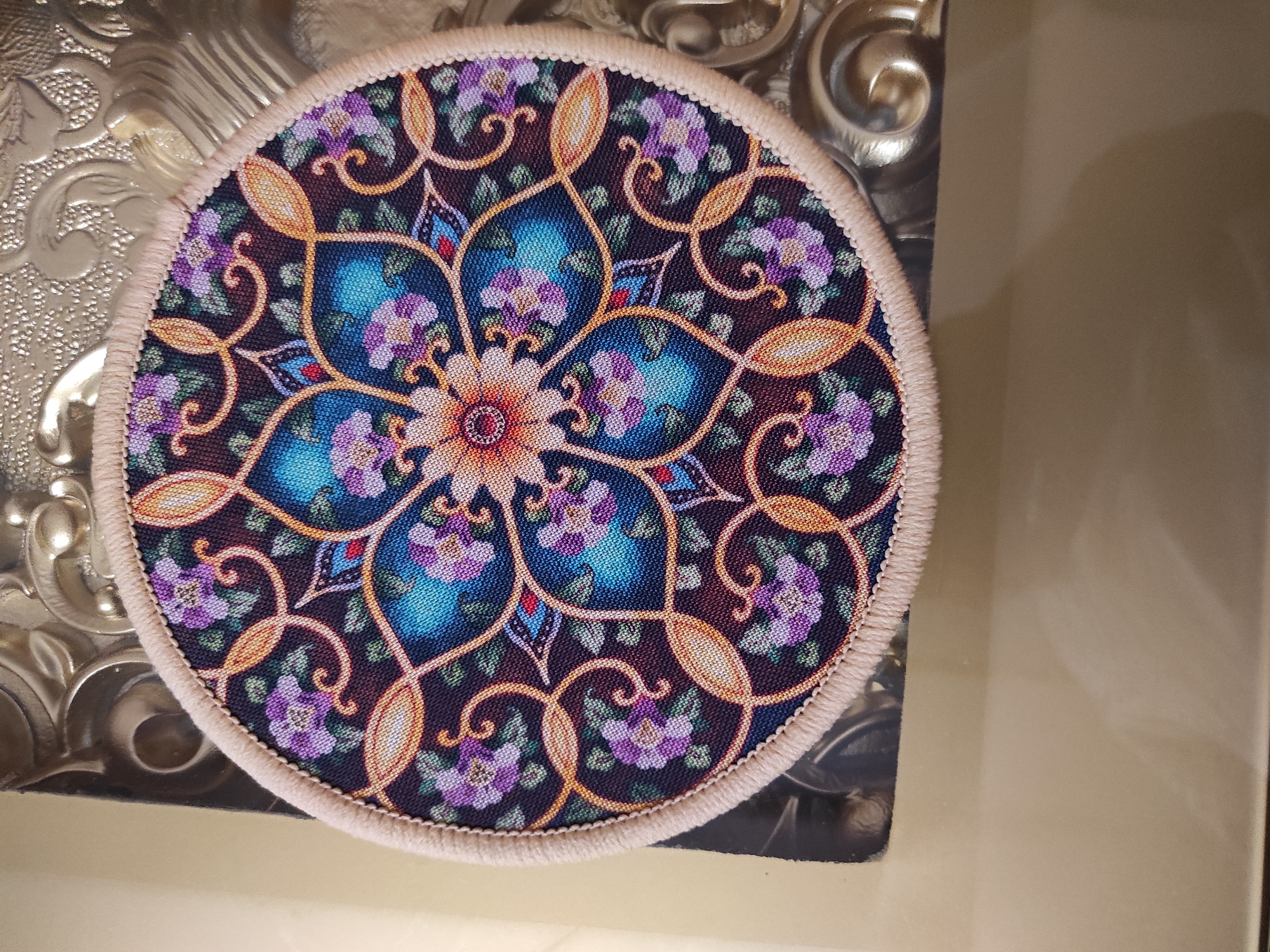 A circular, embroidered coaster with a floral mandala design rests on a textured, metallic surface, likely a decorative tray or tabletop. The coaster features intricate patterns in shades of purple, blue, gold, and red against a dark background. It is trimmed with a light-colored braided edge. The metallic surface appears ornate with swirling, embossed designs. The scene is well-lit, with reflections visible on the metallic surface and a soft, warm ambient light. The background is out of focus, suggesting a shallow depth of field. The coaster is centrally positioned on the metallic surface.