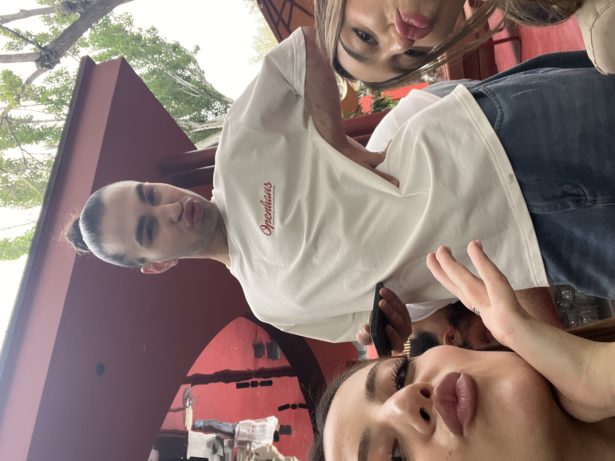 Three people are posing for a selfie under an architectural structure. The person in the white shirt is wearing blue jeans, and the writing "Openhaus" is visible on the shirt. The location appears to be Yerevan, Armenia. There are trees and other architectural elements in the background.