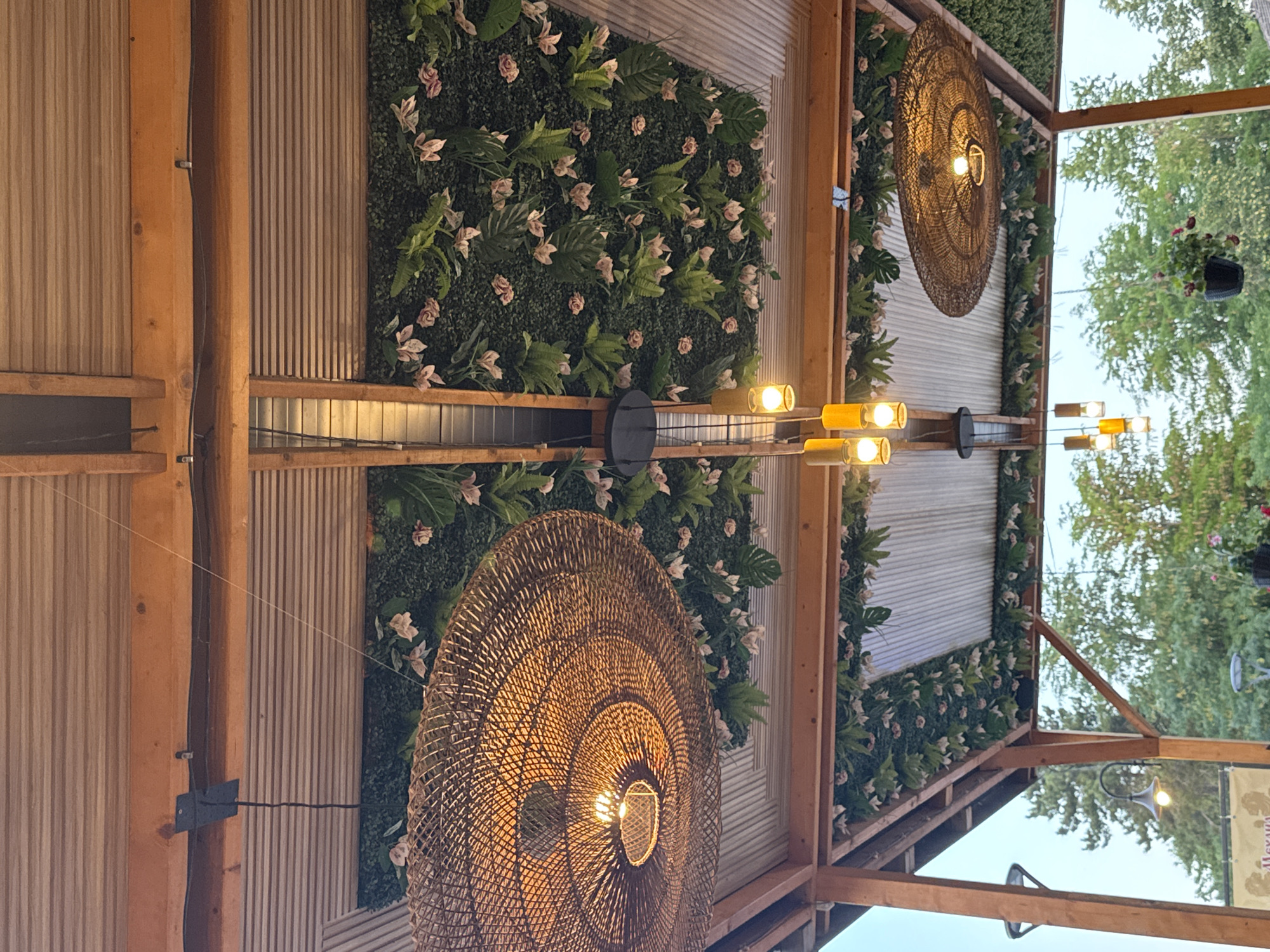 The image shows an outdoor covered area with a wooden pergola structure supporting a corrugated metal roof. The ceiling is adorned with artificial greenery and pink flowers. Two large, woven, circular pendant lights hang from the ceiling, illuminating the space with a warm glow. Several smaller, cylindrical pendant lights are also visible. In the background, trees and sky are visible, suggesting an outdoor setting. The overall impression is of an aesthetically decorated outdoor dining or seating area, likely a restaurant or cafe. The image does not provide explicit information about people, actions, or interactions.