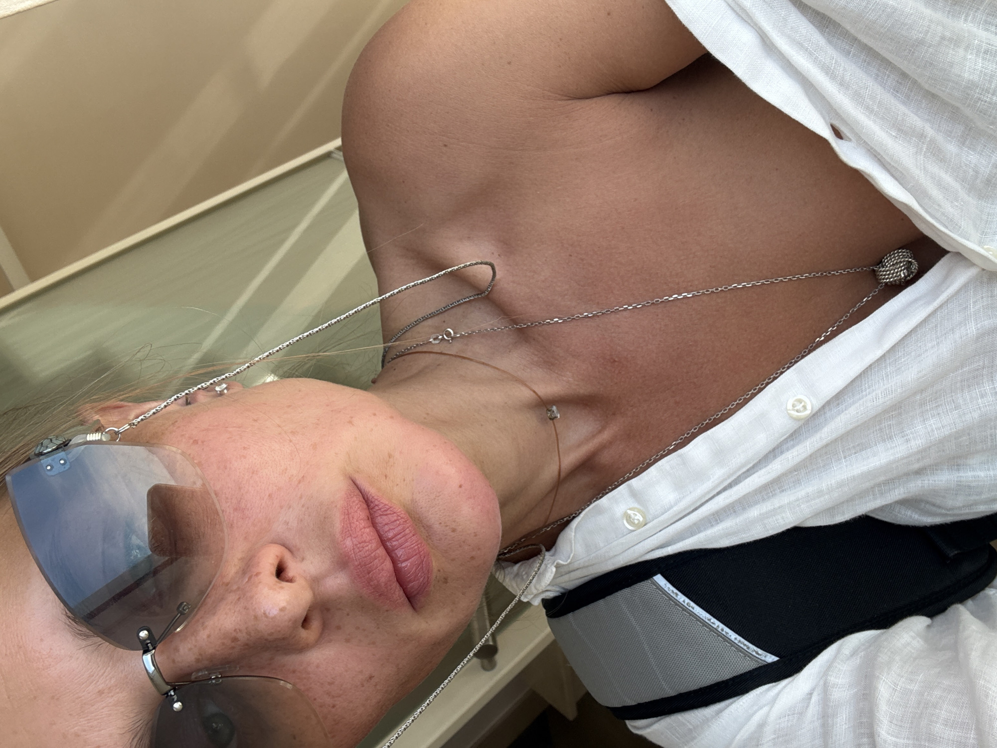 The image shows a close-up, overhead shot of a woman's face and upper torso. She is wearing sunglasses, a white linen shirt, and several silver necklaces. The woman has fair skin with freckles, and her lips are painted with a pink lipstick. She is looking directly at the camera. The background is blurred, but appears to be an interior space with light-colored walls and a window. The scene is likely taken indoors or on a balcony, given the lighting and the presence of what might be a window reflection. It is not possible to determine the location within Budva, Montenegro based on the visual information provided.