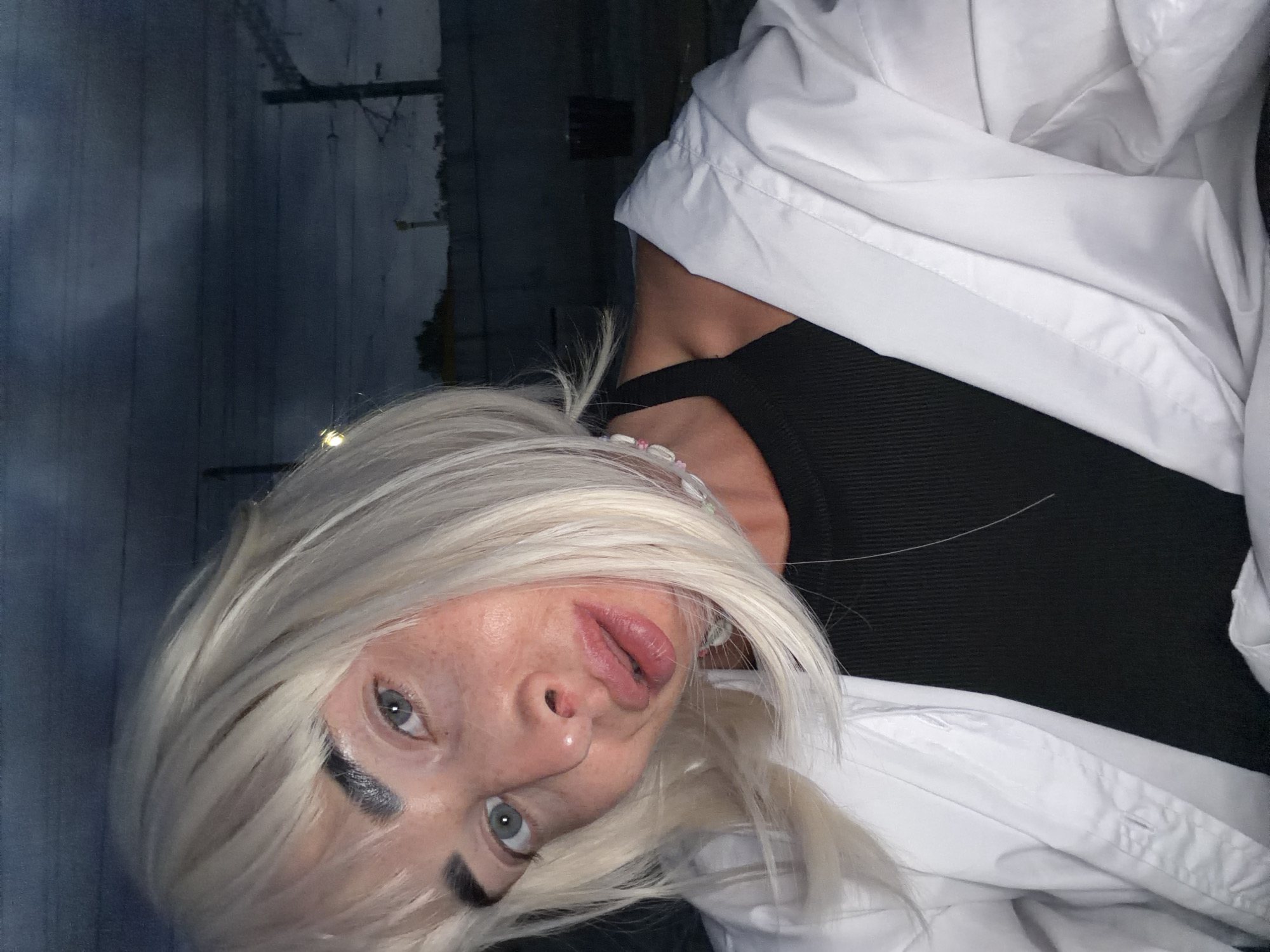 A woman with blonde hair and light blue eyes is pictured in a close-up selfie. She is wearing a black tank top and an open white shirt. A necklace with pink and white beads is visible around her neck. The background appears to be an outdoor urban setting at dusk, with dark clouds and what looks like railway infrastructure. The location is not specified as Sochi, Russia.