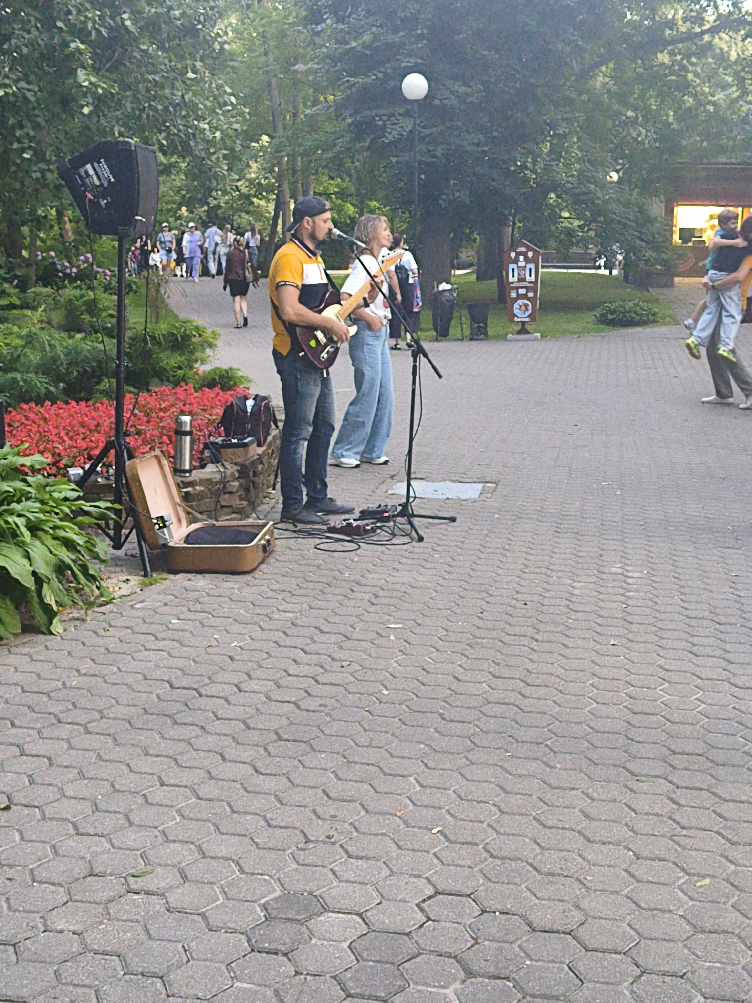 An outdoor scene depicts two musicians performing in a park with a paved walkway. The musicians are standing in the foreground, with one playing an electric guitar and the other singing into a microphone. A PA system speaker and various audio equipment are set up nearby, along with an open vintage suitcase. In the background, other park visitors can be seen walking along the path, and trees and lush vegetation surround the area. A sign is visible to the right, and a building with illuminated windows is in the distance. The park's paved surface consists of interlocking hexagonal stones. This location is the Gomel Central Park in Homyel, Belarus.