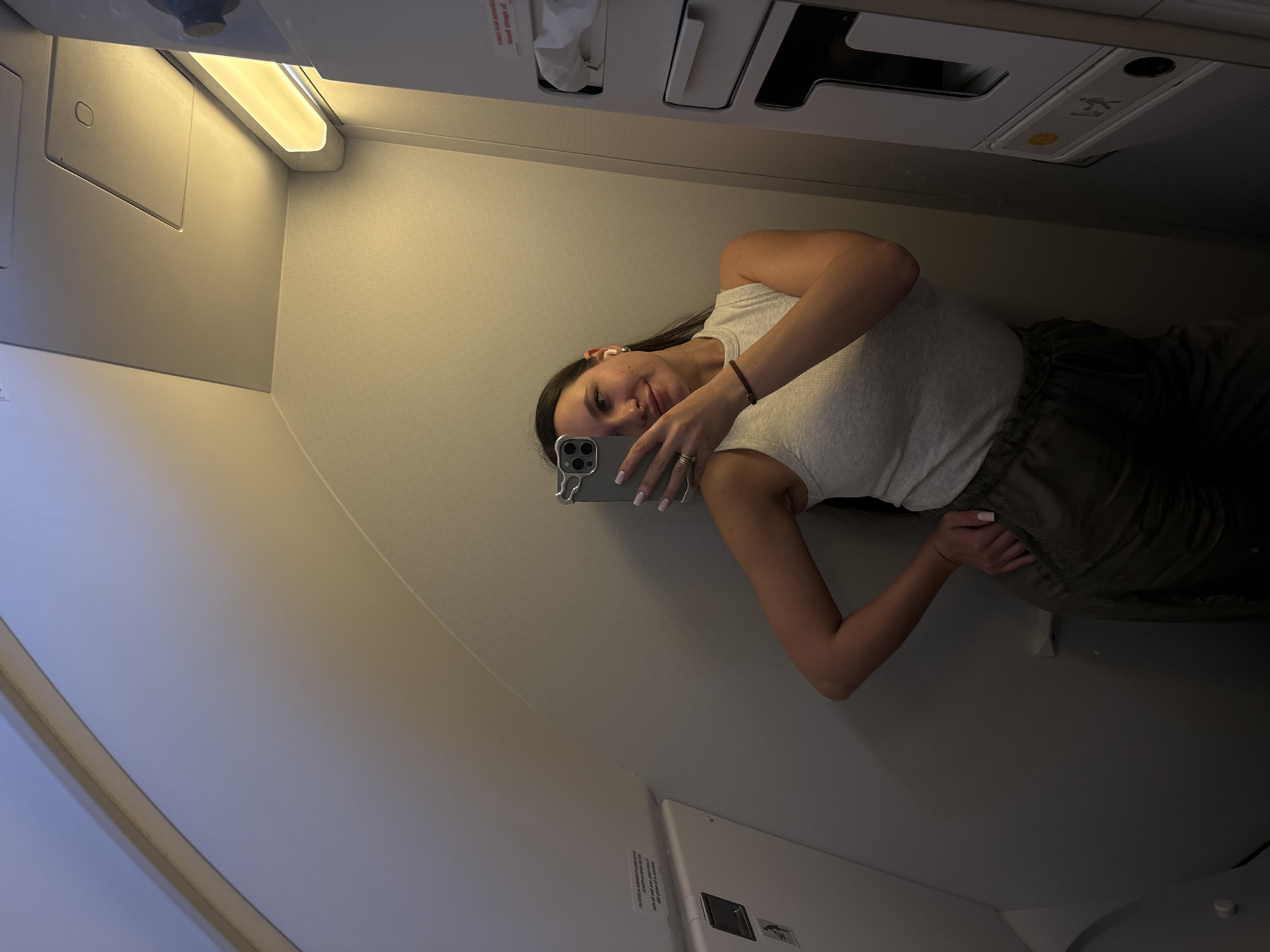 A woman is taking a selfie on an airplane. She is standing in the aisle and holding her phone up to her face. The plane's interior is visible, including the overhead compartments and seats. The location is not discernible as Moscow, Russia.