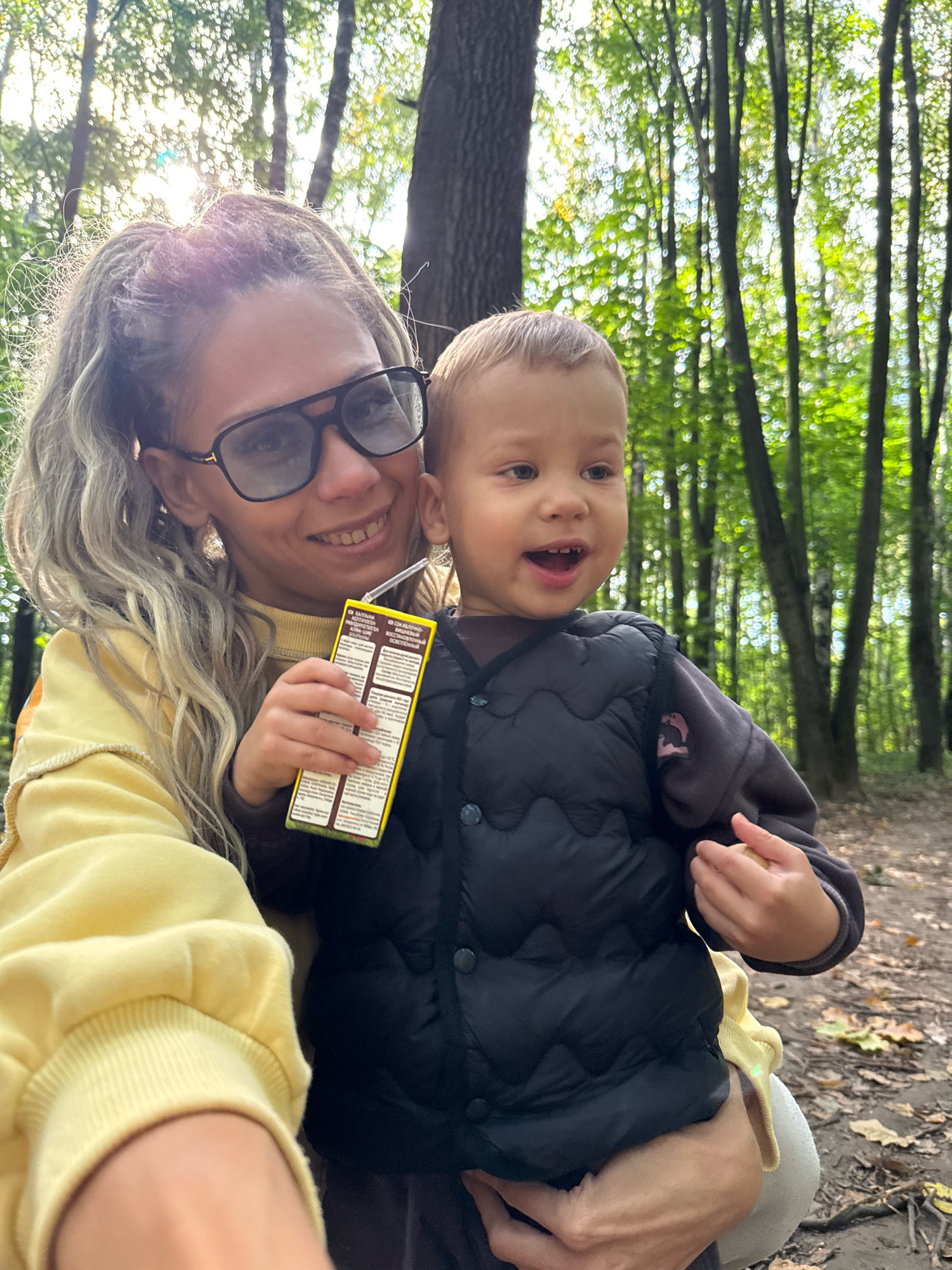 A woman with blonde braided hair and large black glasses holds a young child in her arms. She is wearing a yellow sweatshirt. The child is wearing a dark vest over a long-sleeved shirt. The woman is holding a small juice box with a straw. They are in a forest setting, with trees and fallen leaves visible. The sun is shining through the trees, creating a dappled light effect. The image appears to be a selfie. The specific location in the city of Unknown, Unknown cannot be determined.