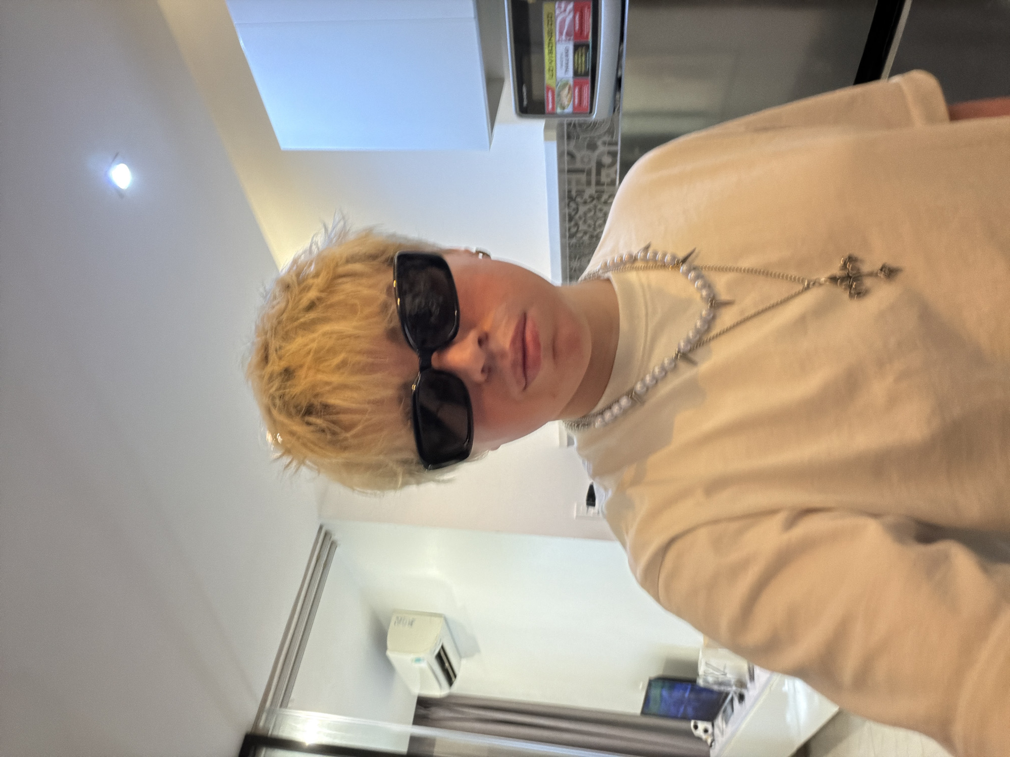A person with short, blond, spiky hair, wearing black rectangular sunglasses and a white t-shirt, is posing for a selfie in what appears to be a modern kitchen area. They are adorned with a pearl necklace and a silver cross pendant. In the background, a white air conditioning unit is mounted on a white wall, a grey curtain hangs to the left, and a stainless steel refrigerator with a visible microwave is to the right. The lighting suggests an indoor environment, possibly in Thủ Đức, Vietnam.