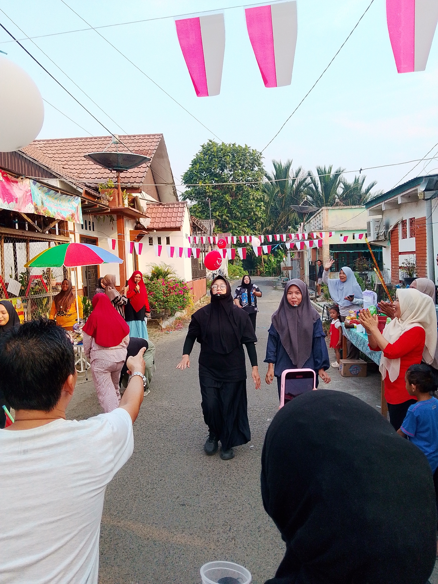 In Barabai, Indonesia, a street is decorated with pink and white banners and red balloons for a festive event. Several people are present, some appearing to participate in a game or activity. A woman in a black hijab and dress is in motion, possibly running or dancing, with a playful expression. To her left, a person in a grey hijab has a piece of candy in their mouth, looking towards the camera. On the right side of the street, a group of women are gathered around a table laden with items, some clapping. A small child in a red dress stands near the table, while another child in a blue shirt observes the scene. In the foreground, a person wearing a white t-shirt is visible from behind, facing away from the camera. The background shows houses with tiled roofs and trees, characteristic of a residential area in a town or city. The lighting suggests it is daytime, possibly late afternoon.