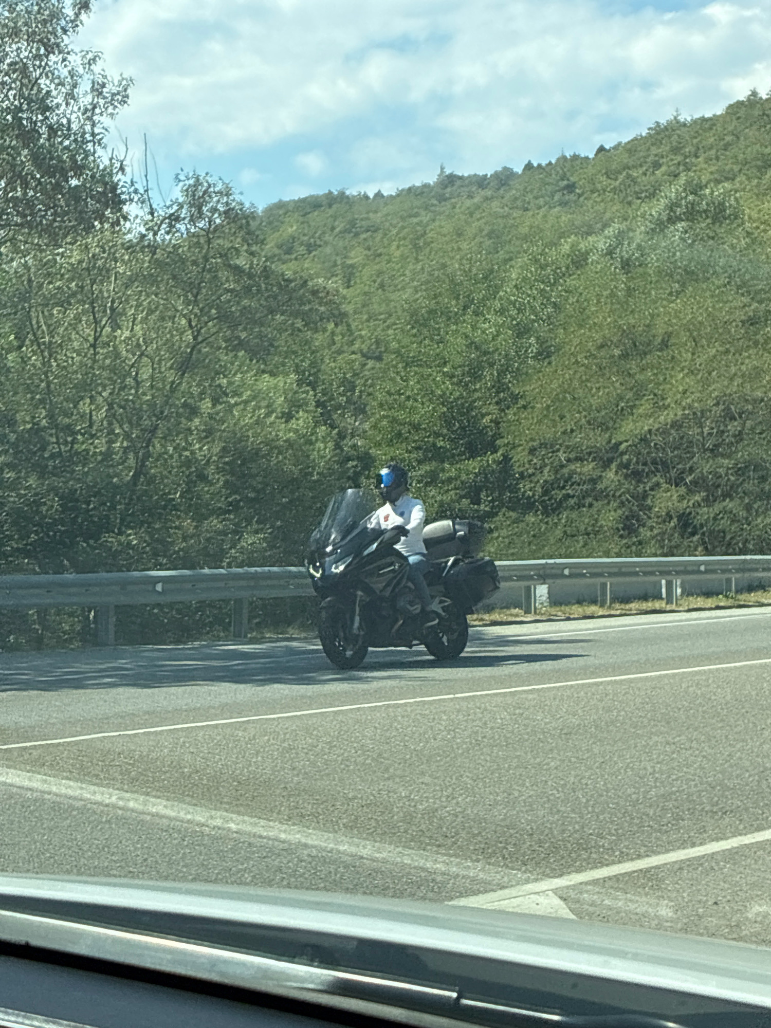A motorcyclist wearing a white shirt and a blue helmet rides a black touring motorcycle on a highway. The motorcycle has two side cases and a large windshield. The highway is bordered by a metal guardrail and a dense forest with green trees covering rolling hills in the background. The sky is partly cloudy with a bright blue hue. The perspective suggests the photo was taken from another vehicle on the highway. The image does not contain any specific landmarks or visual cues to pinpoint the exact location within Shirokaya Schel, Russia, other than the general environment of a highway passing through a forested, hilly region.