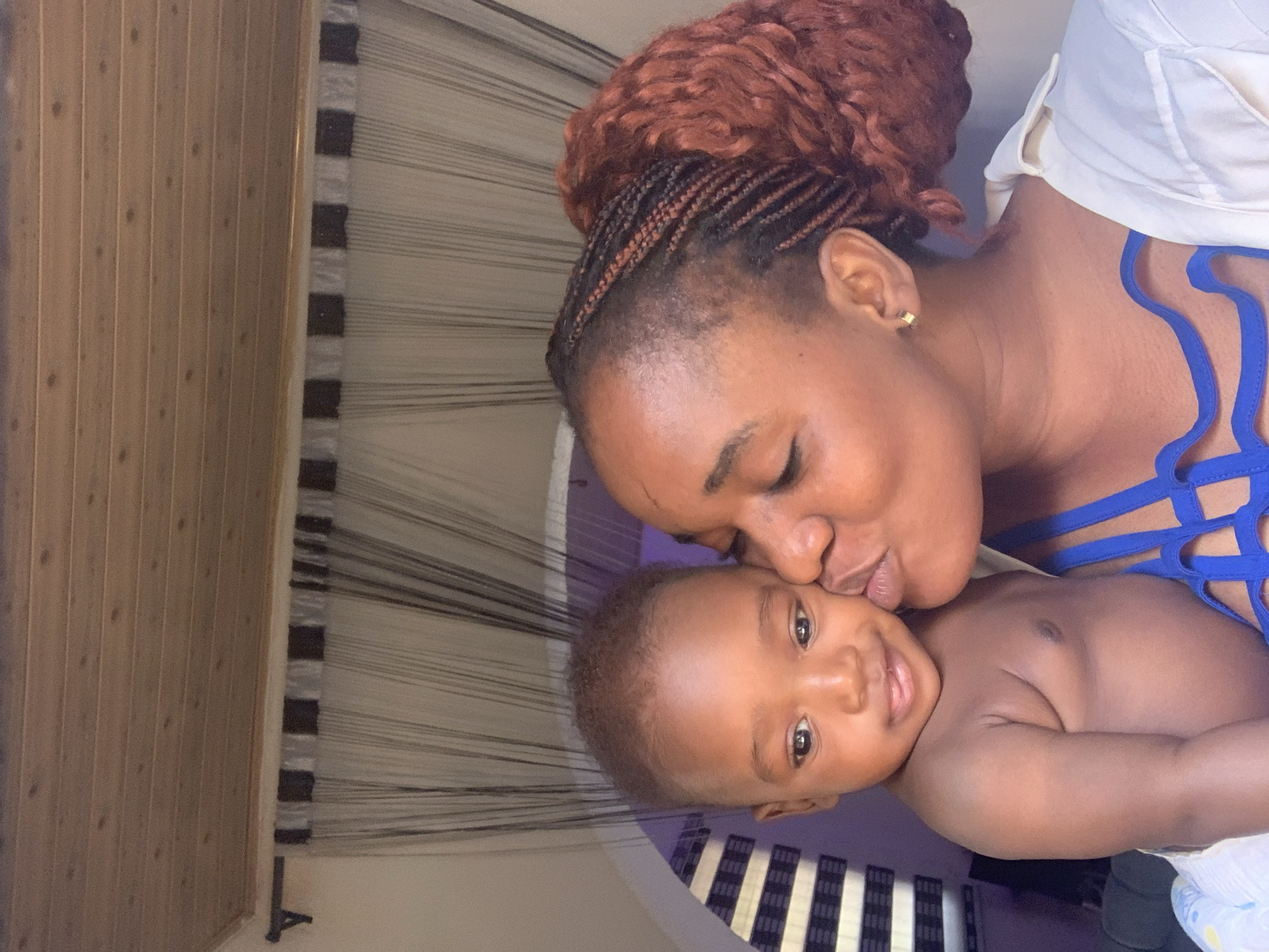 In a close-up, vertical shot, a mother lovingly kisses her baby on the cheek. The scene captures a tender moment between the two subjects, who are positioned in the foreground.
The woman, likely the mother, has dark skin and wears her hair in reddish-brown braids styled into a bun, with some cornrows visible on the side of her head. She has a small golden hoop earring on her right ear. She is wearing a blue top with intricate, strappy patterns, and a portion of a white garment is visible over her right shoulder. Her eyes are closed as she leans in to kiss the baby.
The baby, appearing to be an infant or toddler, has dark skin and short, dark hair. The baby is shirtless, revealing a smooth chest, and wears a light-colored diaper with a subtle blue pattern. The baby is looking directly at the viewer with a slight, content smile, and bright, dark eyes.
The background consists of several elements. To the left, there's a light brown wooden paneled wall. Behind and to the right of the subjects, a distinctive curtain or room divider with numerous thin, dark strings against a lighter background is visible, creating a striped effect. Some parts of this divider also feature wider horizontal bands in black and white or light grey. Further back, a plain purple wall is visible behind the baby's head.
The overall mood of the image is warm and affectionate, highlighting the bond between mother and child. The lighting appears soft, suggesting an indoor setting. No visible text is present in the image. The location context provided is Barakin Akawo, Nigeria.